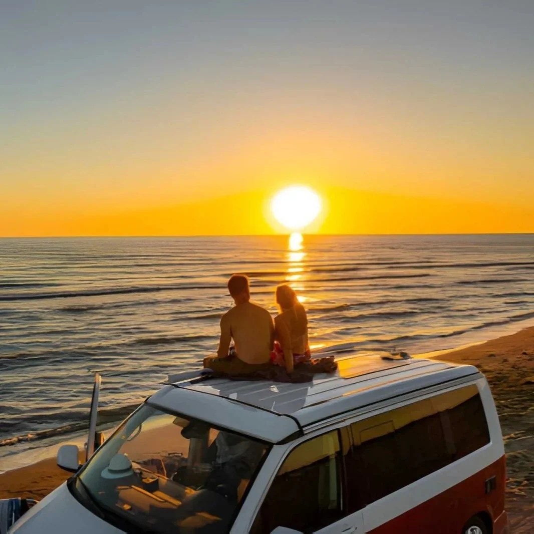 Couple sitting on top of a white camper van on a sandy beach watching a golden sunset over the sea during a road trip in Albania.
