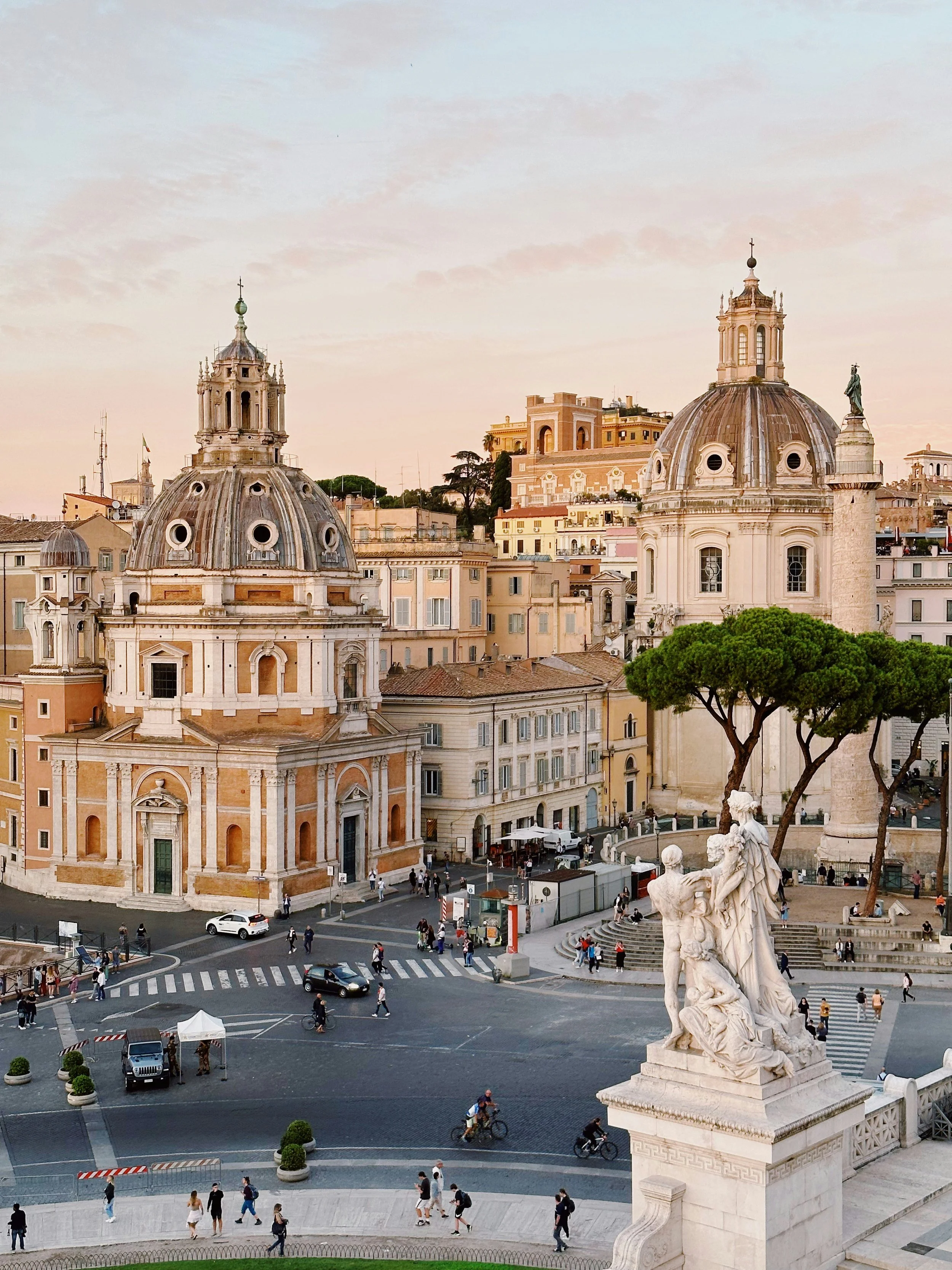 A panoramic view of Rome, Italy, featuring the twin Baroque churches of Santa Maria di Loreto and the Church of the Most Holy Name of Mary, with Trajan’s Column rising between them, seen from the terrace of the Vittoriano.
