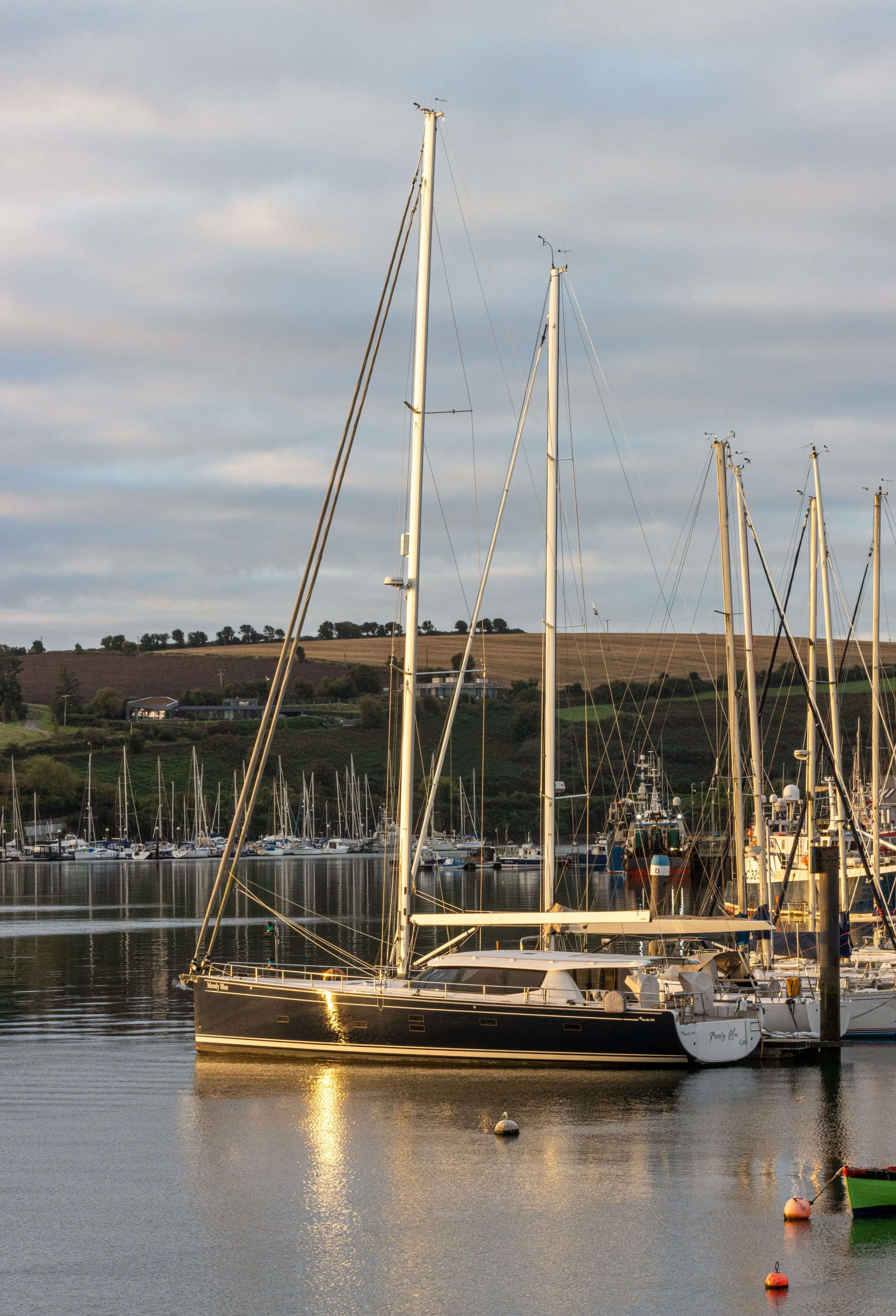 Kinsale Harbour, County Cork, Ireland, with sailboats moored against a backdrop of rolling hills at sunset.