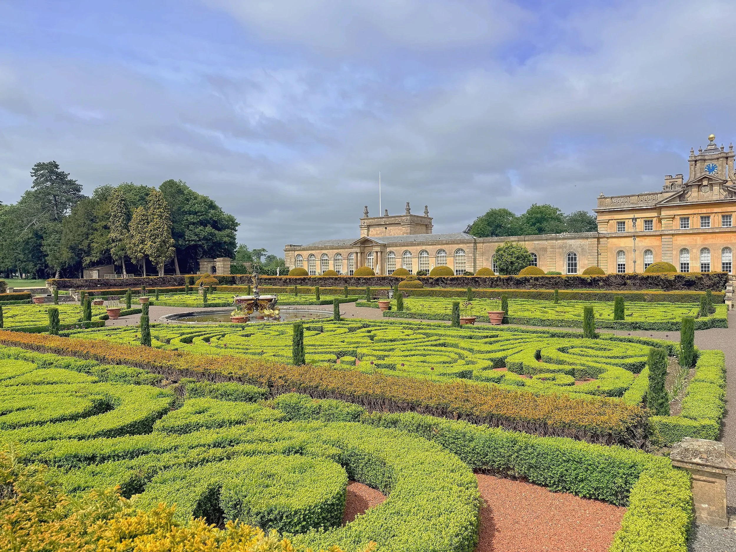 Blenheim Palace in Woodstock, Oxfordshire, England, features grand palace architecture overlooking formal gardens with neatly trimmed parterres and gravel pathways.