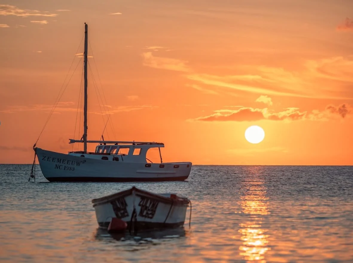 Sunset over Boca Sami, Curaçao, with sailboats floating on calm waters beneath a colorful evening sky.
