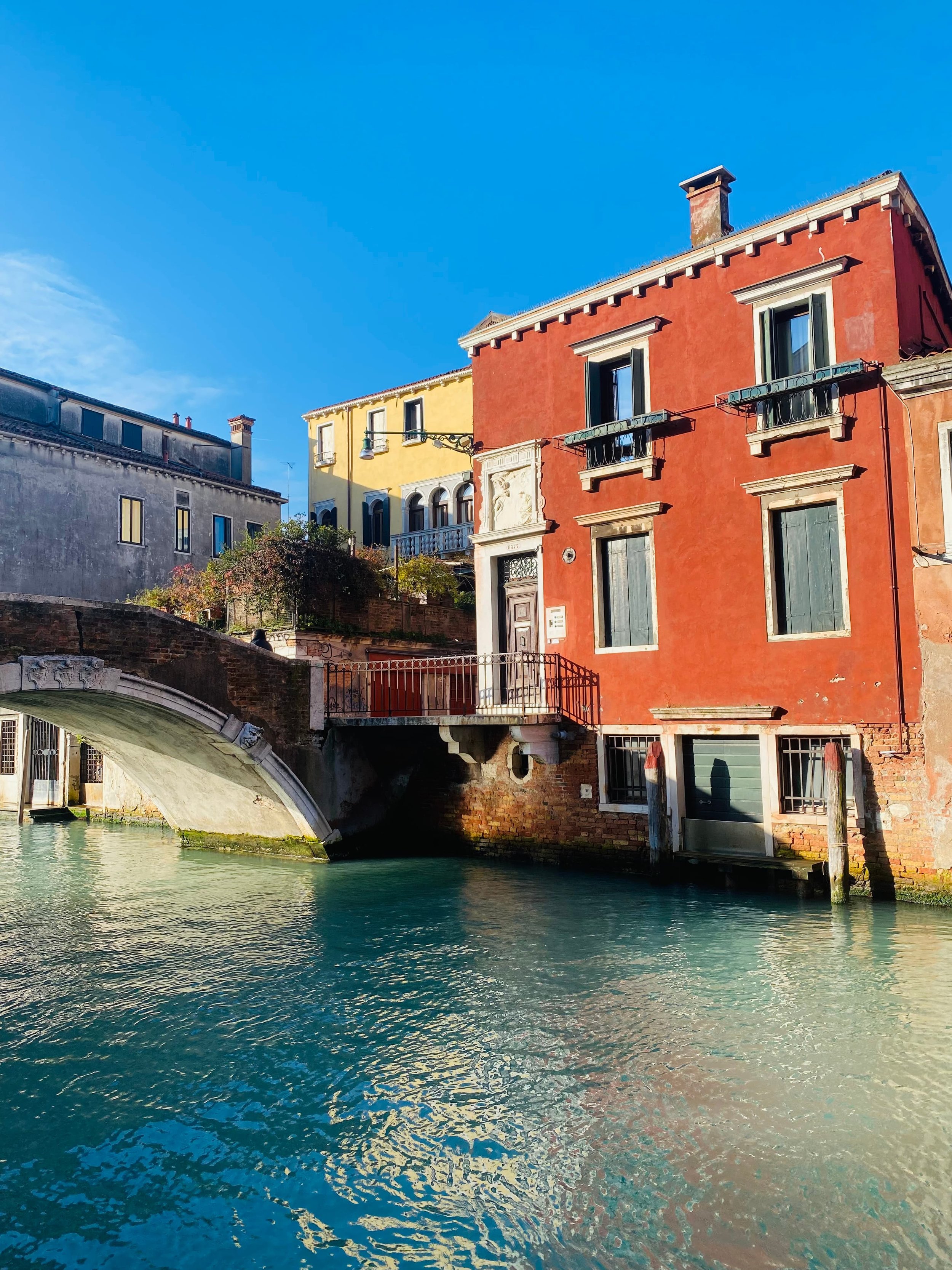 A scenic canal in Venice, Italy, with a historic red building beside a stone footbridge crossing the water, showcasing the city’s charming architecture and picturesque waterways.