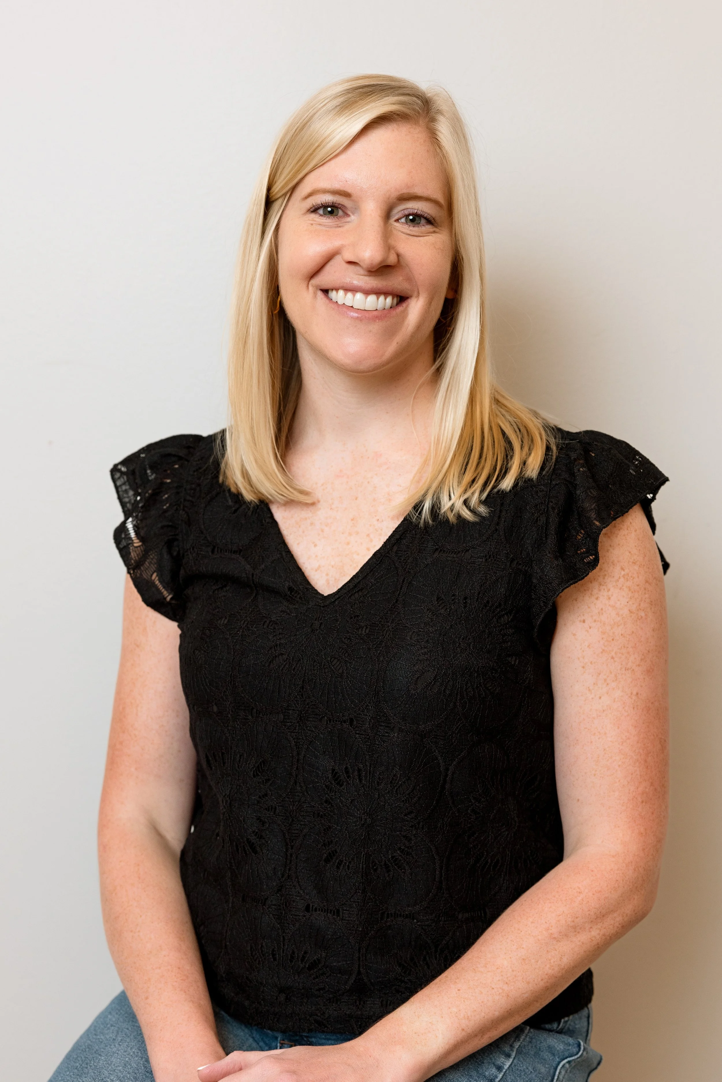 A woman with blonde hair wearing a black V-neck shirt and blue jeans, sitting against a plain light background, smiling at the camera.