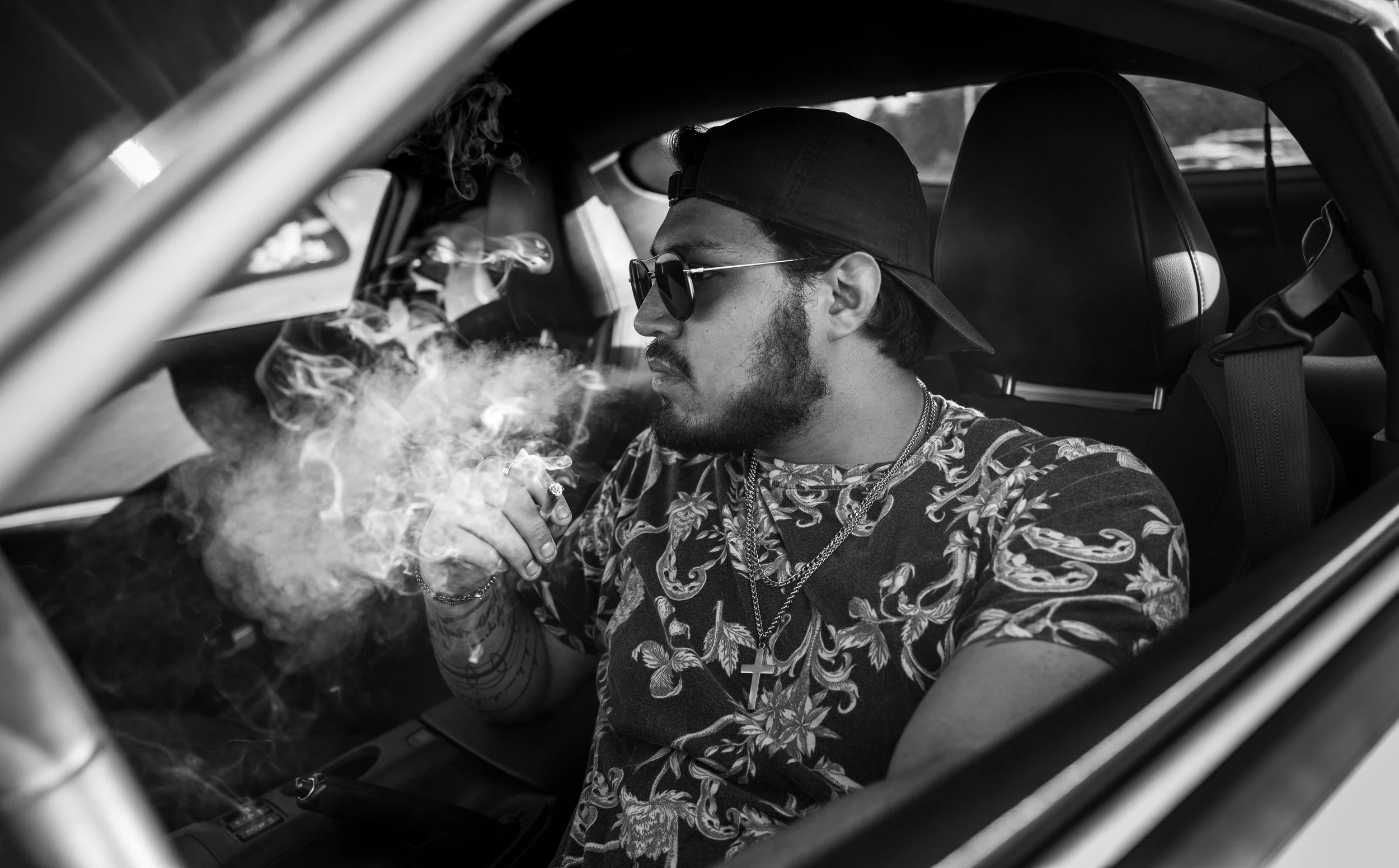 A man with a beard and sunglasses sitting inside a car, wearing a cap backward and a floral shirt, smoking a cigarette.