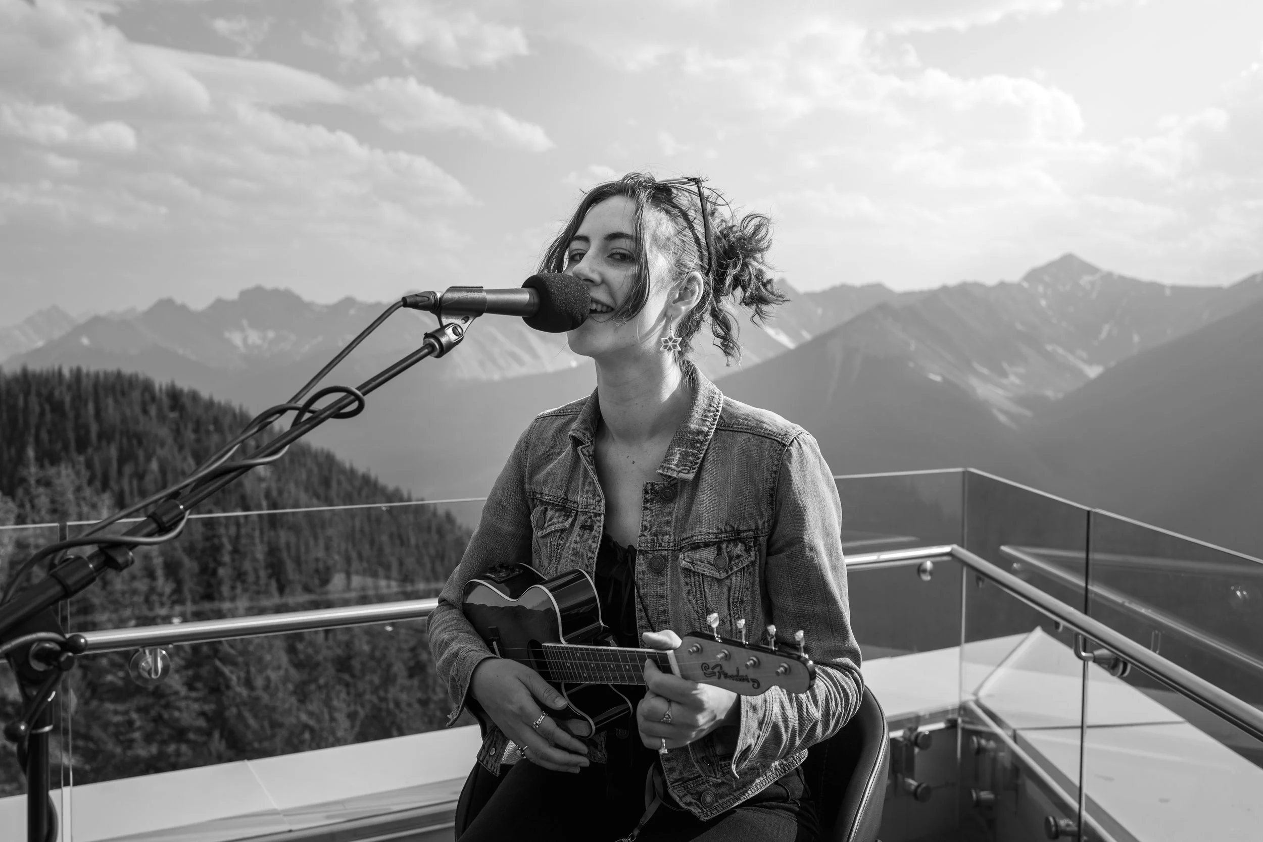 A woman with curly hair wearing a denim jacket and earrings, playing a small guitar and singing into a microphone on a rooftop with mountain scenery in the background.