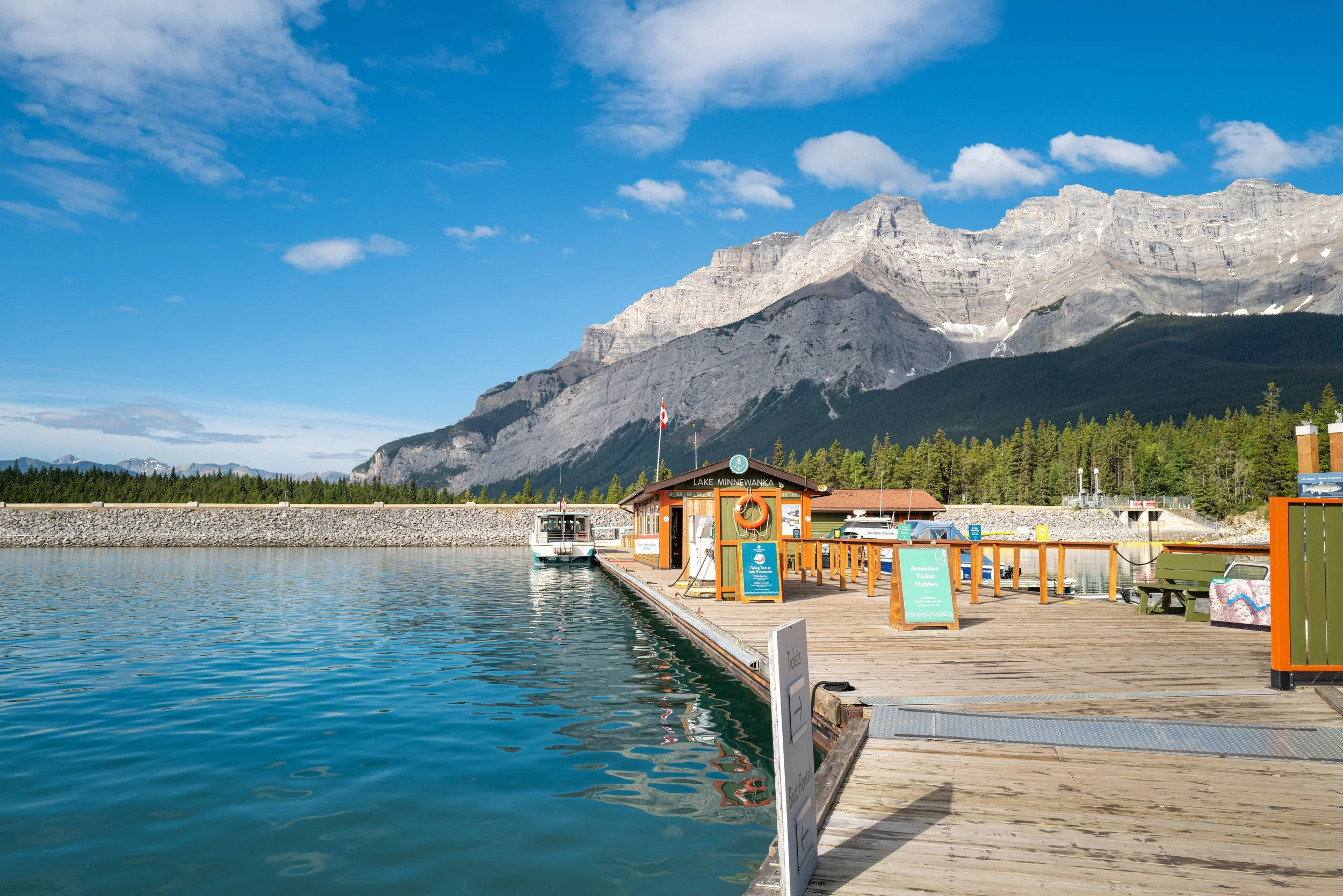 A lakeside dock with boats, a small building, and signs, with tall mountains and a partly cloudy blue sky in the background.