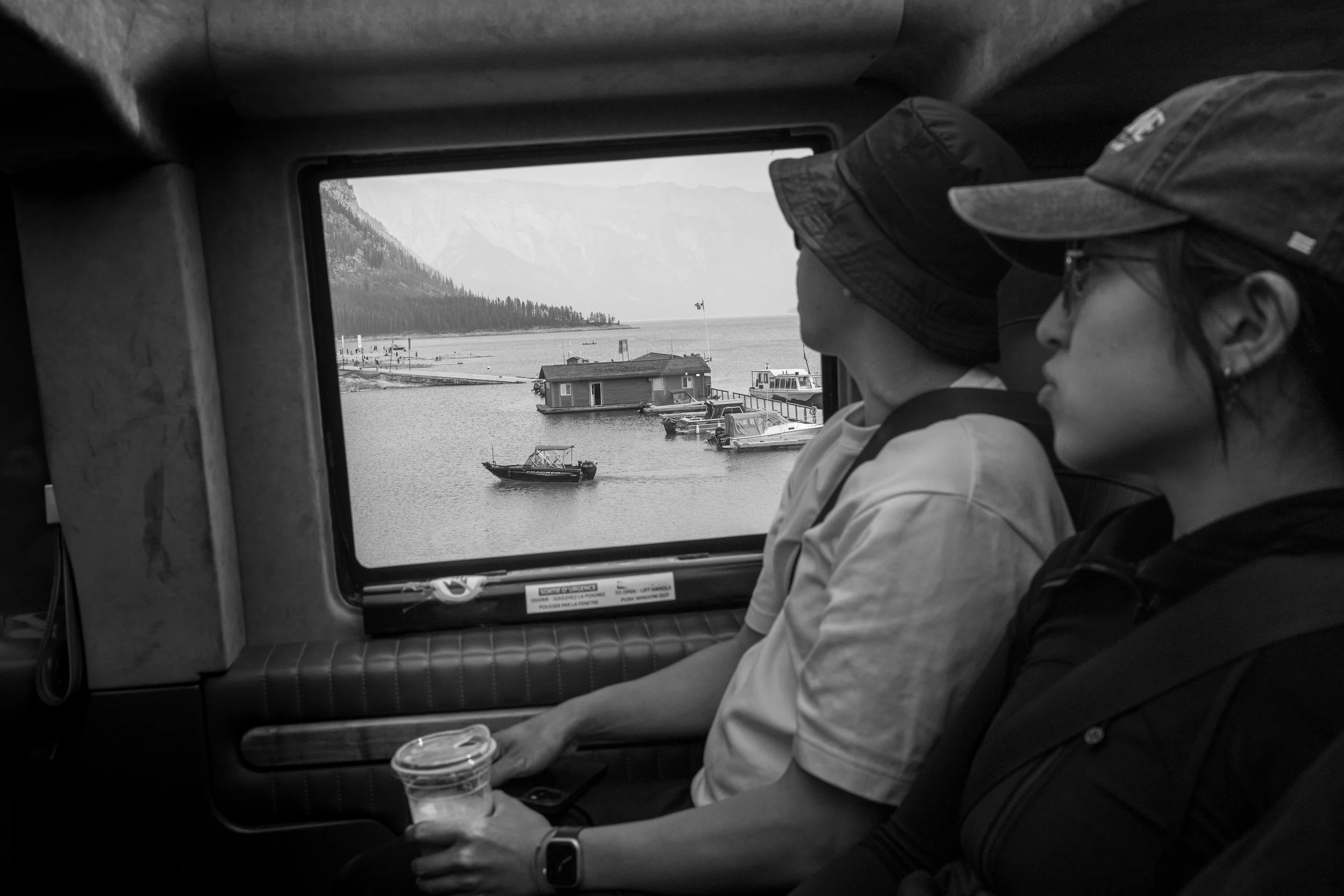 Two women wearing hats sitting inside a vehicle, looking out the window at a body of water, boats, and mountains in the background.