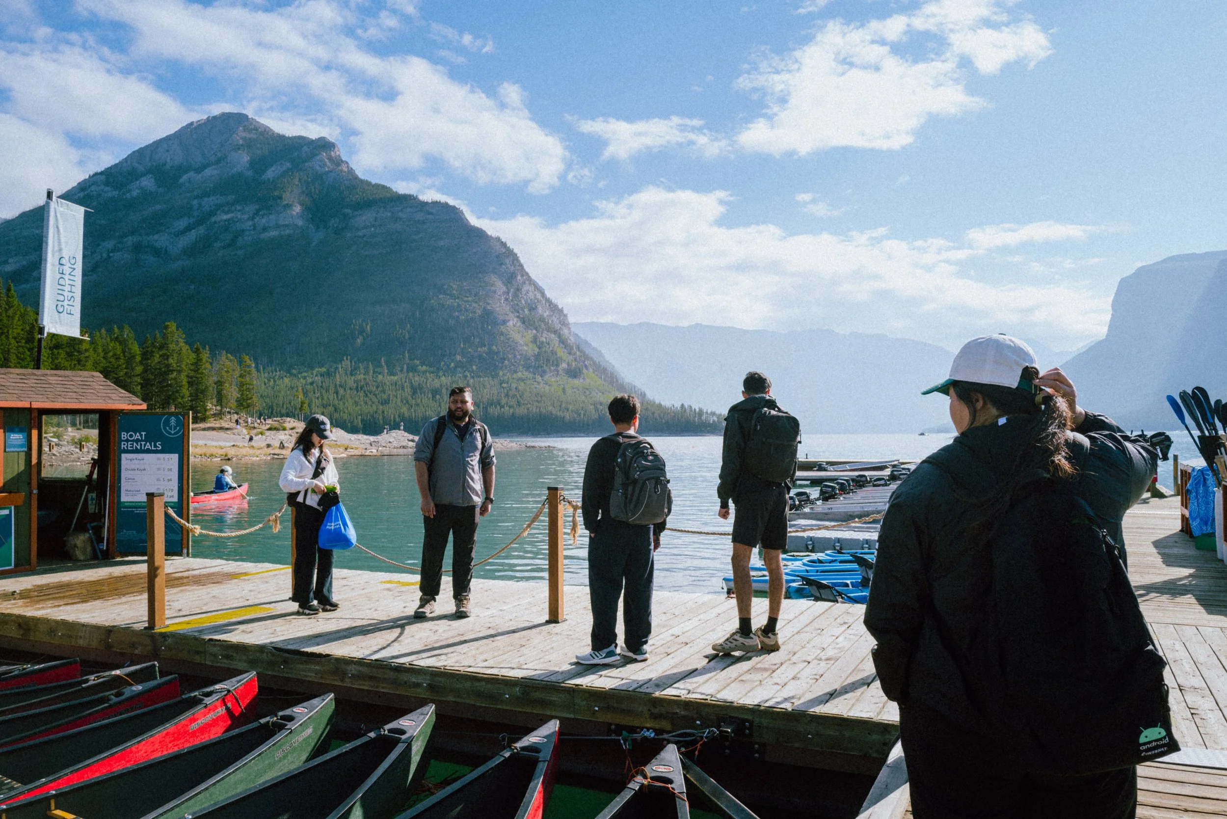 People preparing for kayaking at a lakeside dock with mountains and blue sky in the background.