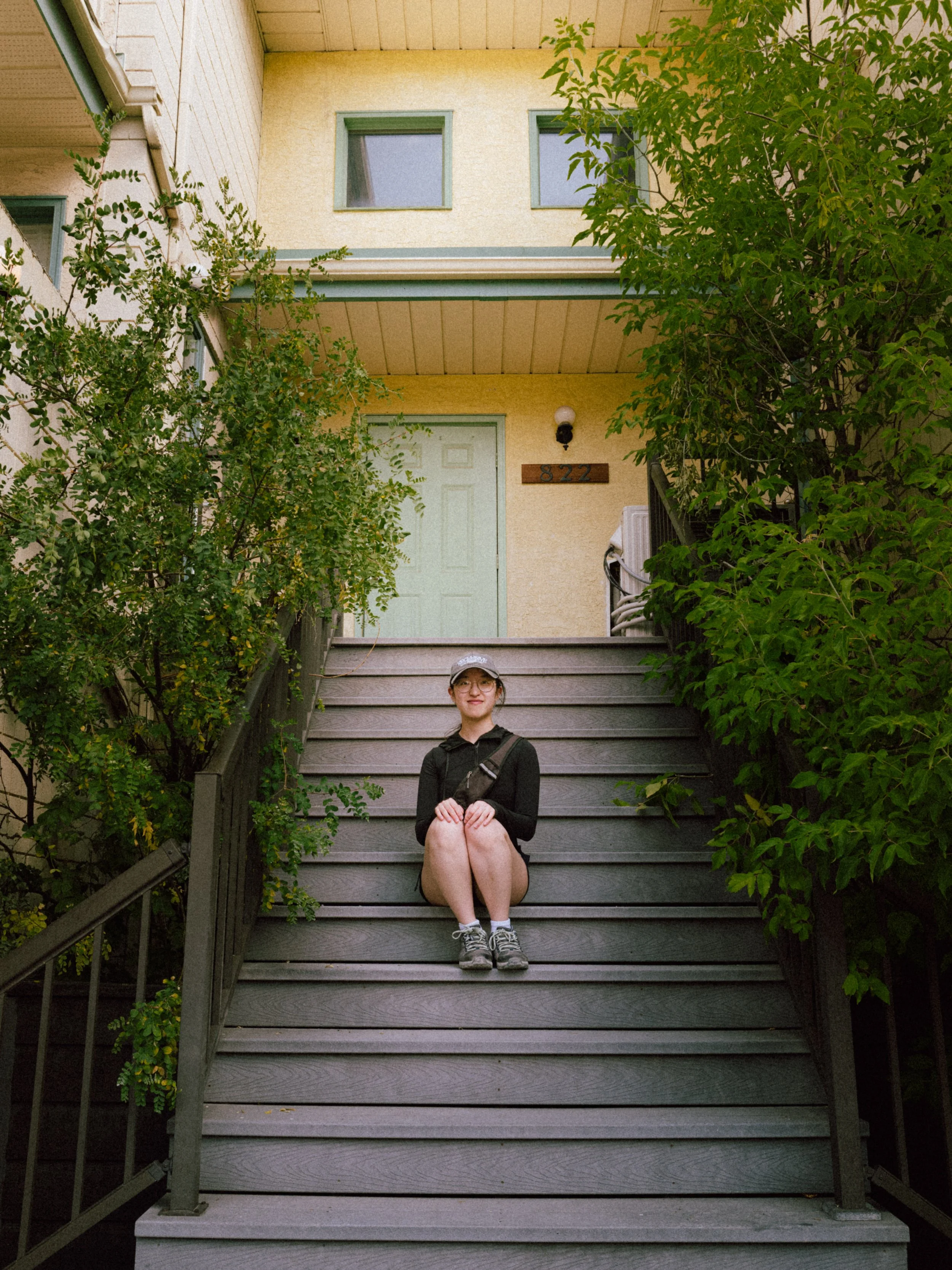 A young woman sitting on outdoor stairs with greenery on both sides, in front of a yellow house with two windows, a door, and house number 822.