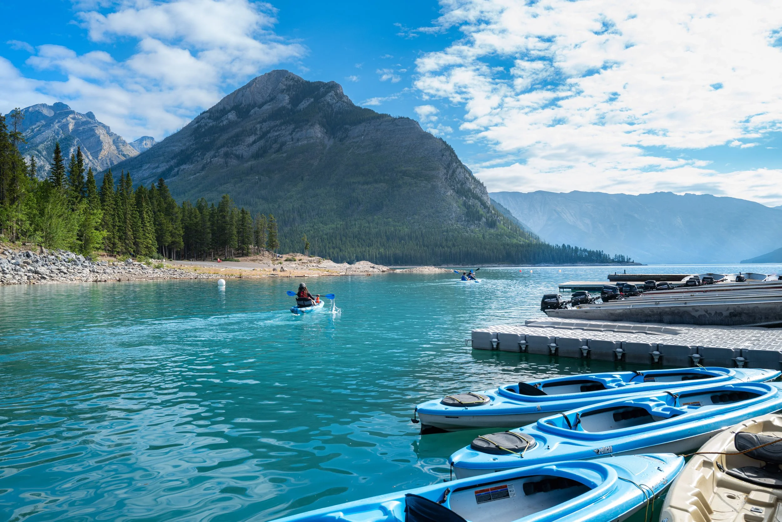 Kayakers paddle on a turquoise lake with a backdrop of mountains, pine trees, a blue sky with scattered clouds, and boats docked along a floating dock.
