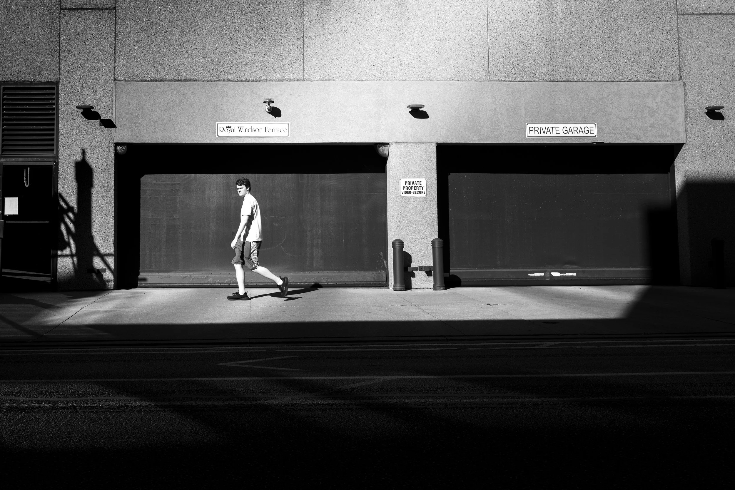 A black and white photo of a building with a garage entrance, a young man in a white shirt and shorts walking in front of it, and signs indicating private property and a private garage.