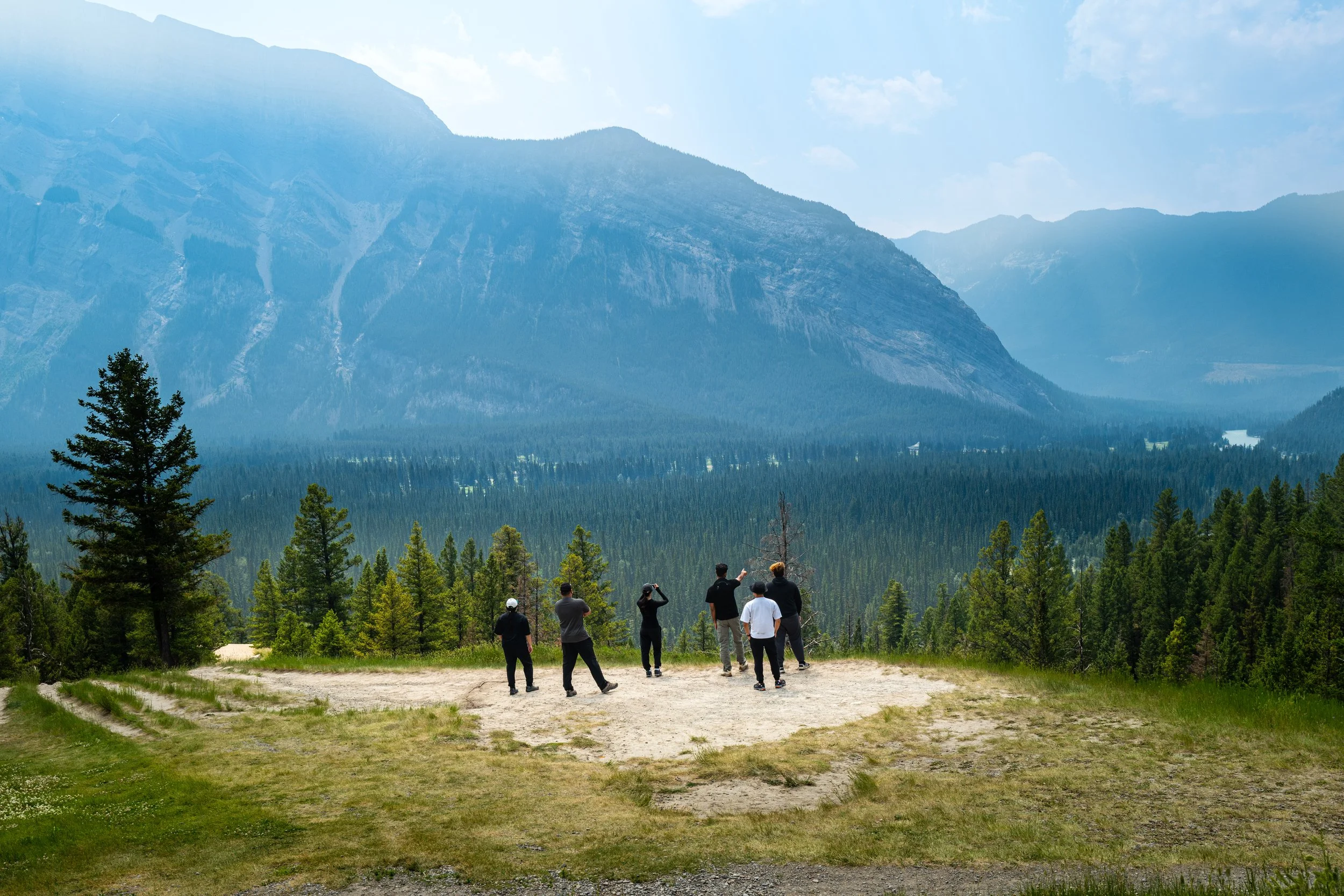 A group of six people standing on a grassy clearing and looking at a mountain range in the distance. The mountains are tall and rugged, with a few patches of snow near the peaks. The sky is partly cloudy with some clouds covering the top of the mount