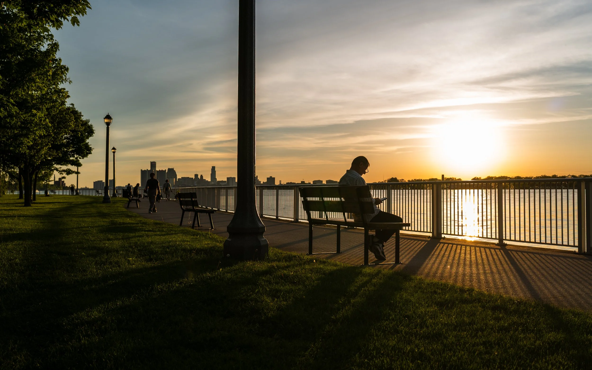 A person sitting on a park bench by a river during sunset, with a city skyline in the background and a few other people walking along the path.