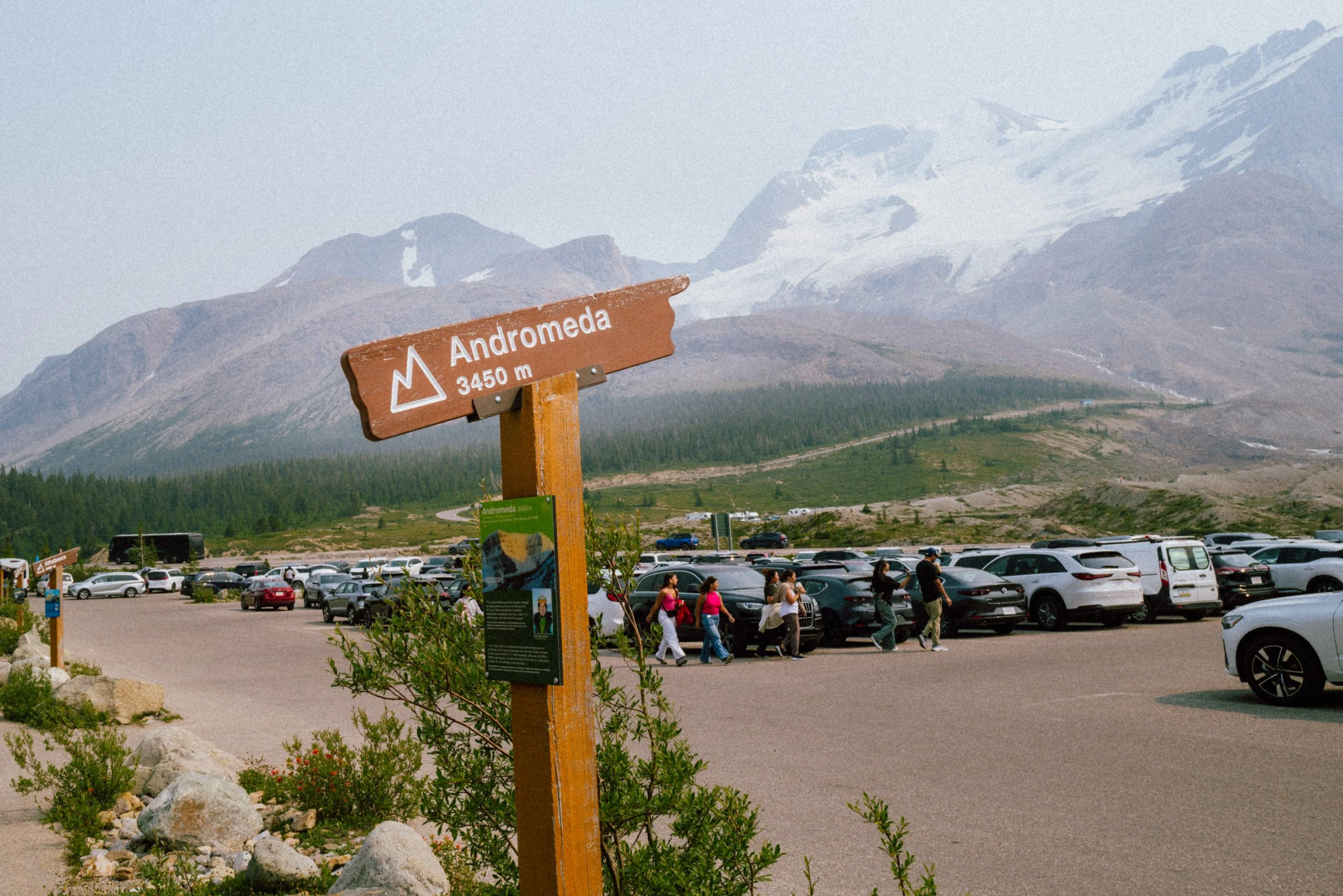 Mountain landscape with a signpost that reads 'Andromeda 3450 meters'; a parking lot filled with cars and people walking; snow-covered mountains in the background.