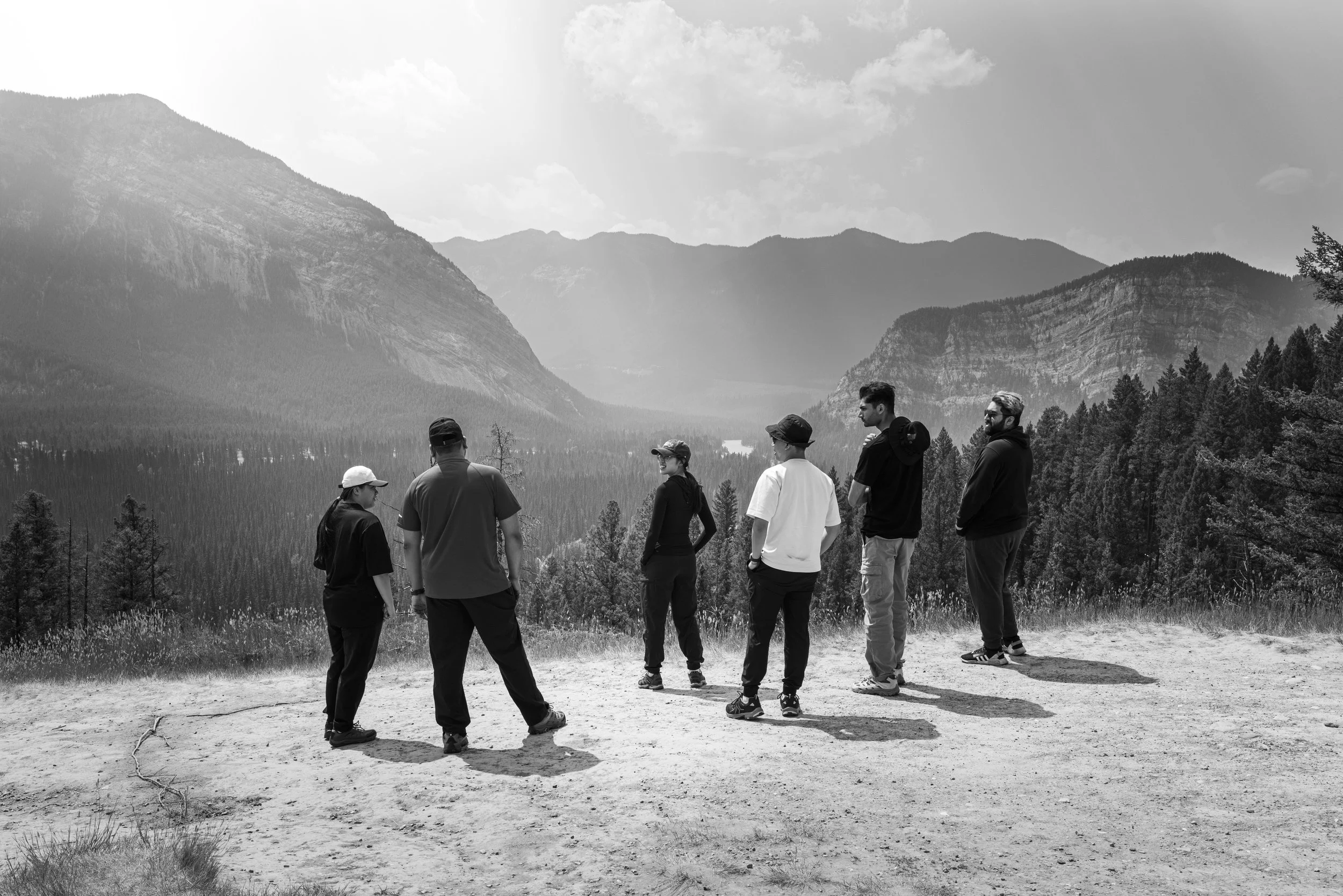 A group of seven people stands on a dirt trail in a valley surrounded by tall mountains and pine trees. The sky has some clouds and the image is in black and white.