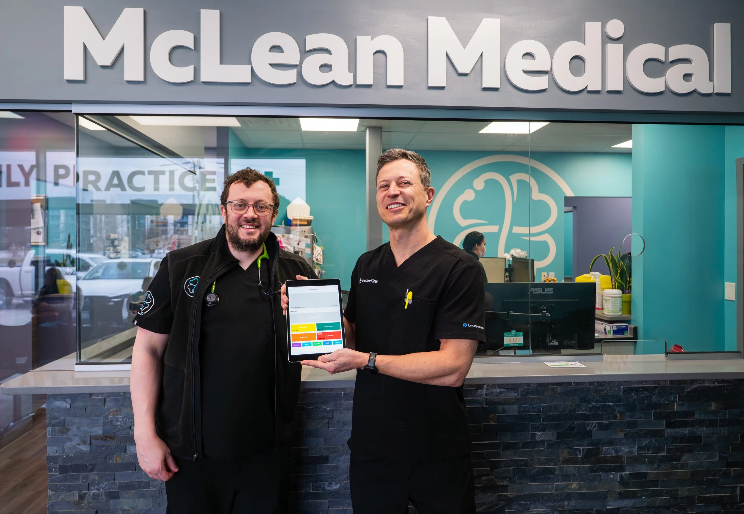 Two men standing in front of McLean Medical sign, smiling, one holding a tablet with a colorful schedule, inside a medical office.