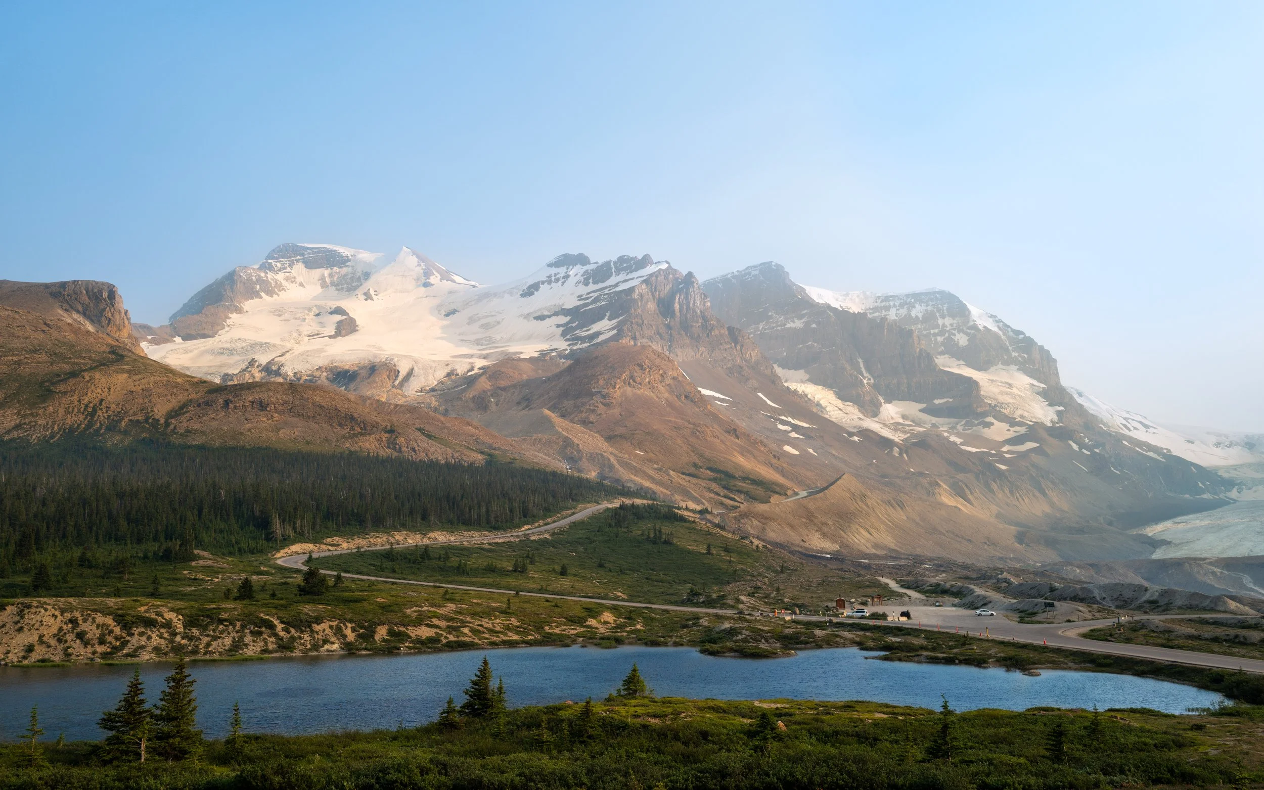 Snow-capped mountains with a forested foreground, a lake, and a winding road with cars.