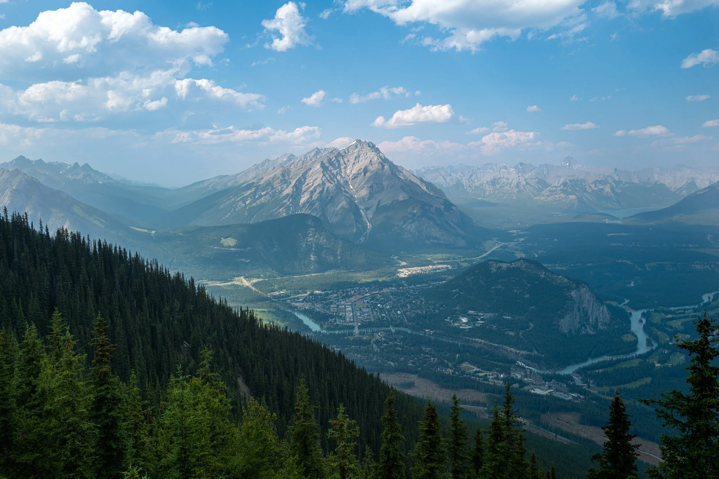 Scenic view of mountain range with green forests in the foreground, a river winding through a valley, and a town at the base of the mountains under a partly cloudy sky.