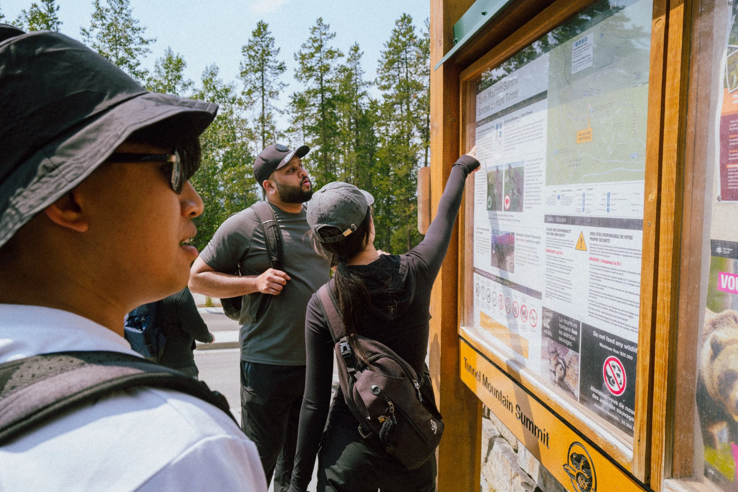 Group of people looking at an informational signboard outdoors surrounded by trees, with some groups pointing and reading the details.