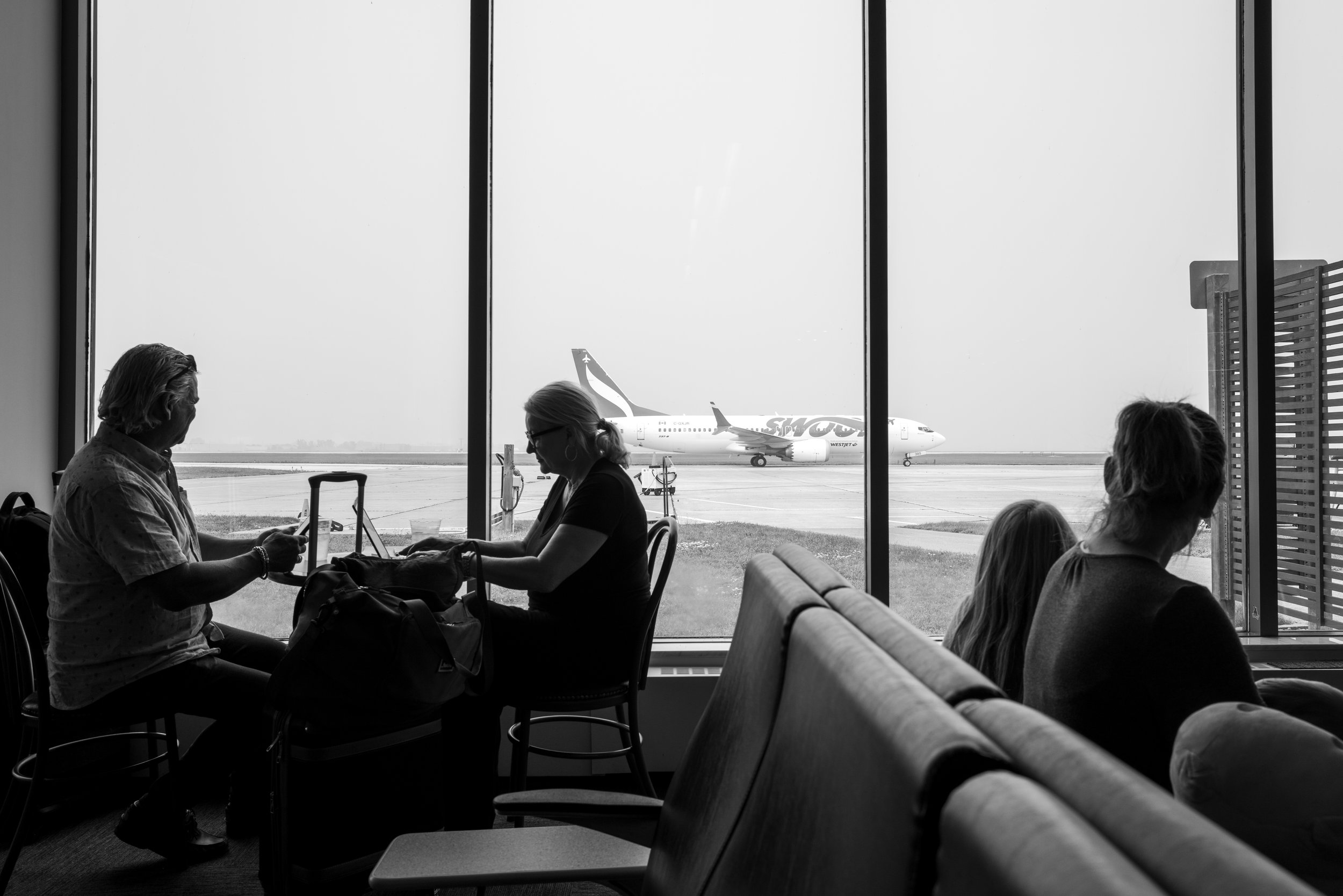 People sitting inside an airport terminal waiting area with a view of an airplane through large windows.