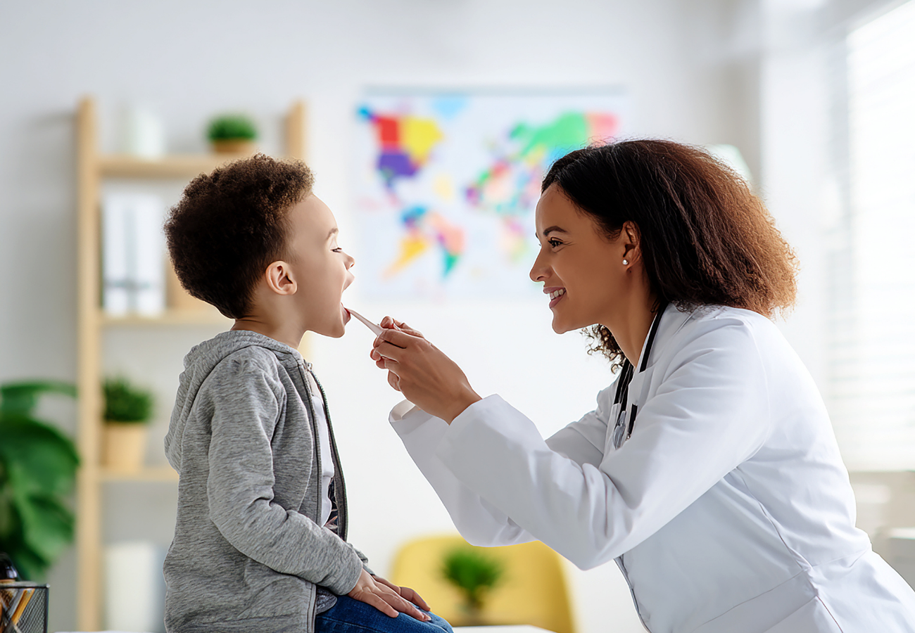 A female doctor examining a young boy's throat with a tongue depressor in a medical office.