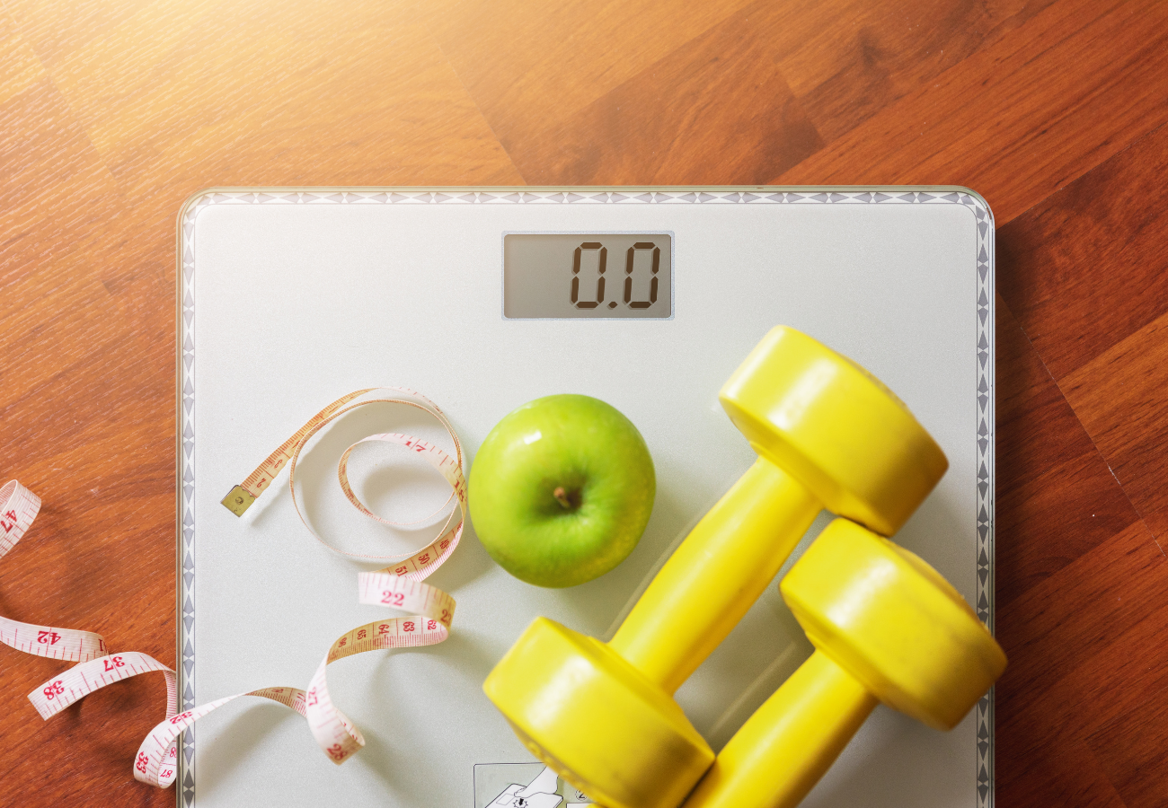 A digital kitchen scale with a green apple, yellow dumbbells, and a measuring tape on it, placed on a wooden surface.