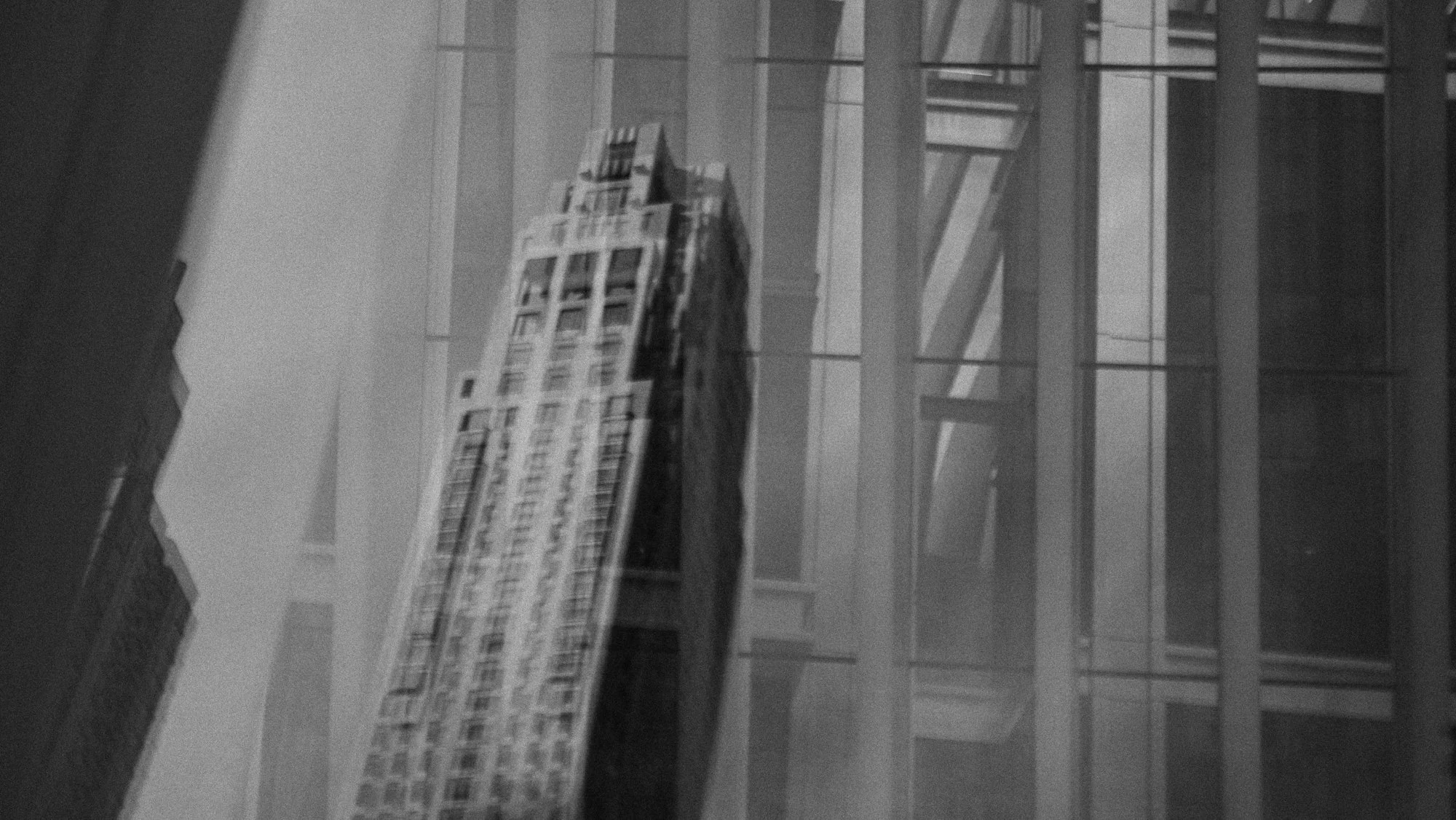 Black and white photo of a tall modern skyscraper in an urban cityscape, surrounded by other buildings and glass structures.