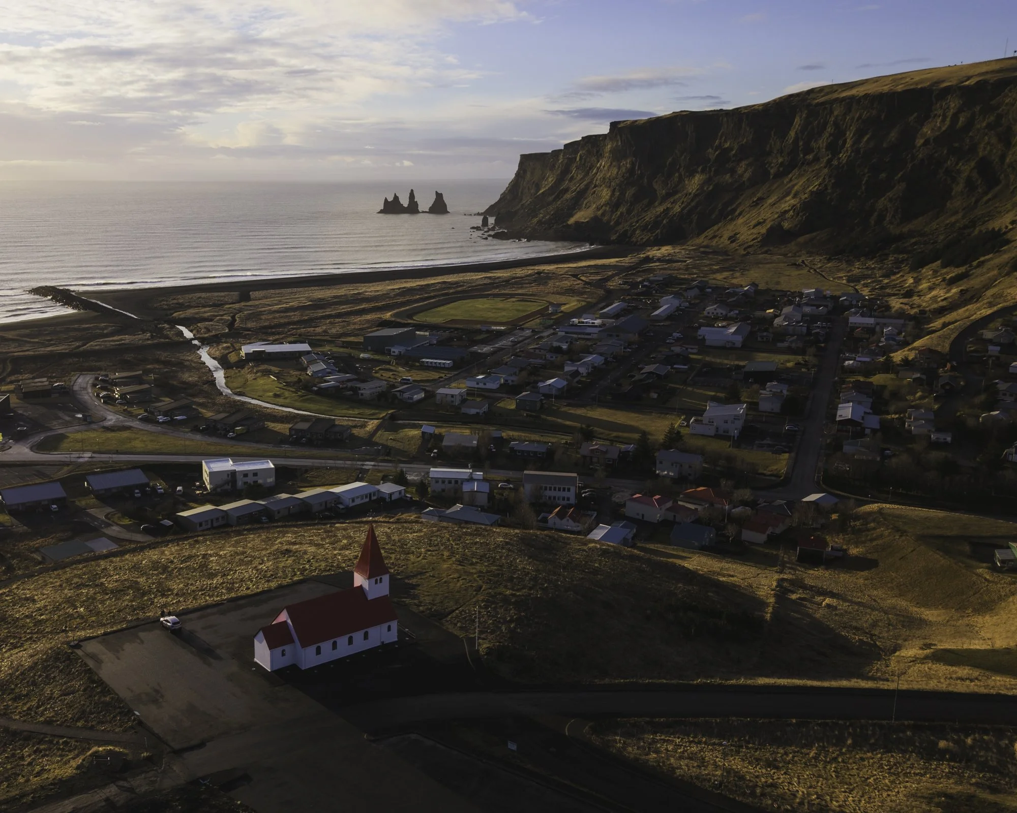 A coastal town on a hillside with a white church with a red roof in the foreground, houses, a track, and cliffs in the background near the ocean with rock formations.