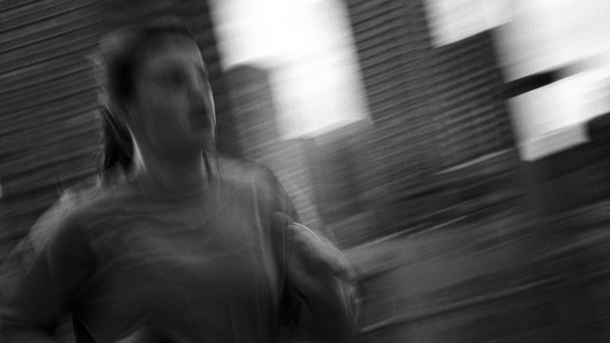A woman in motion, walking past a corrugated metal wall, captured with a blurred, black-and-white effect