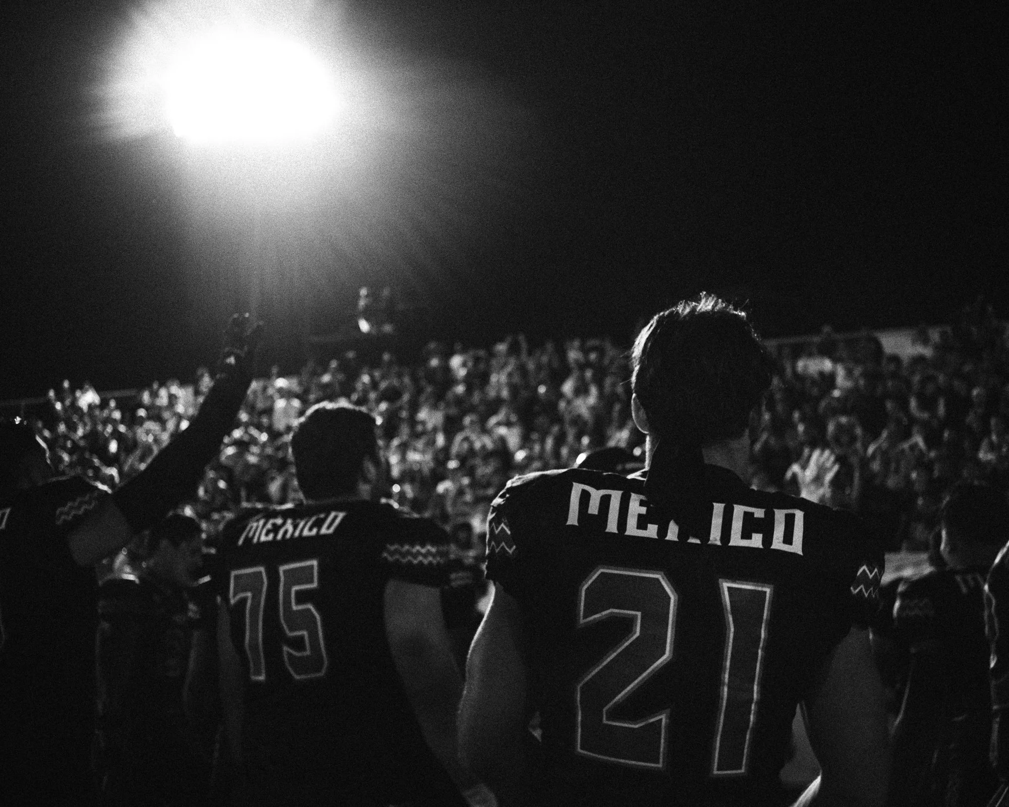 Black and white photo of football players in helmets and jerseys, with one showing the name 'MEXICO' and the number '21', facing a crowd during a night game under bright stadium lights.