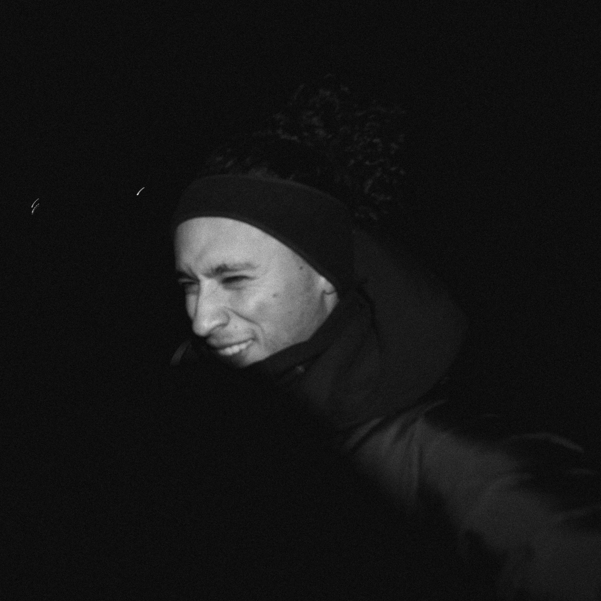 A person outdoors at night, wearing a beanie and a jacket, taking a selfie with a slight squint and a playful expression.