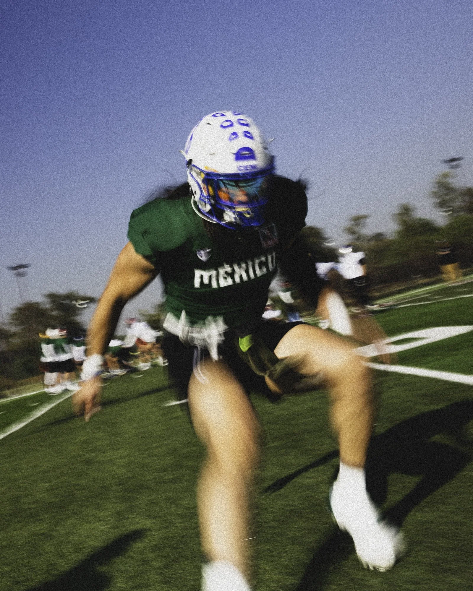 American football player in green uniform with 'Mexico' on it, wearing a helmet and visor, jumping or falling on the field during a game.