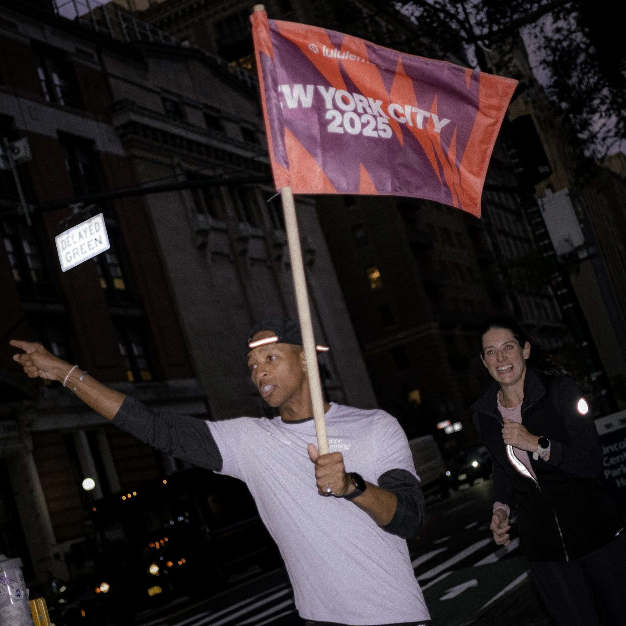 Two people running in a city street during a race, with one holding a flag that reads 'New York City 2025' and a woman smiling behind, at dusk.