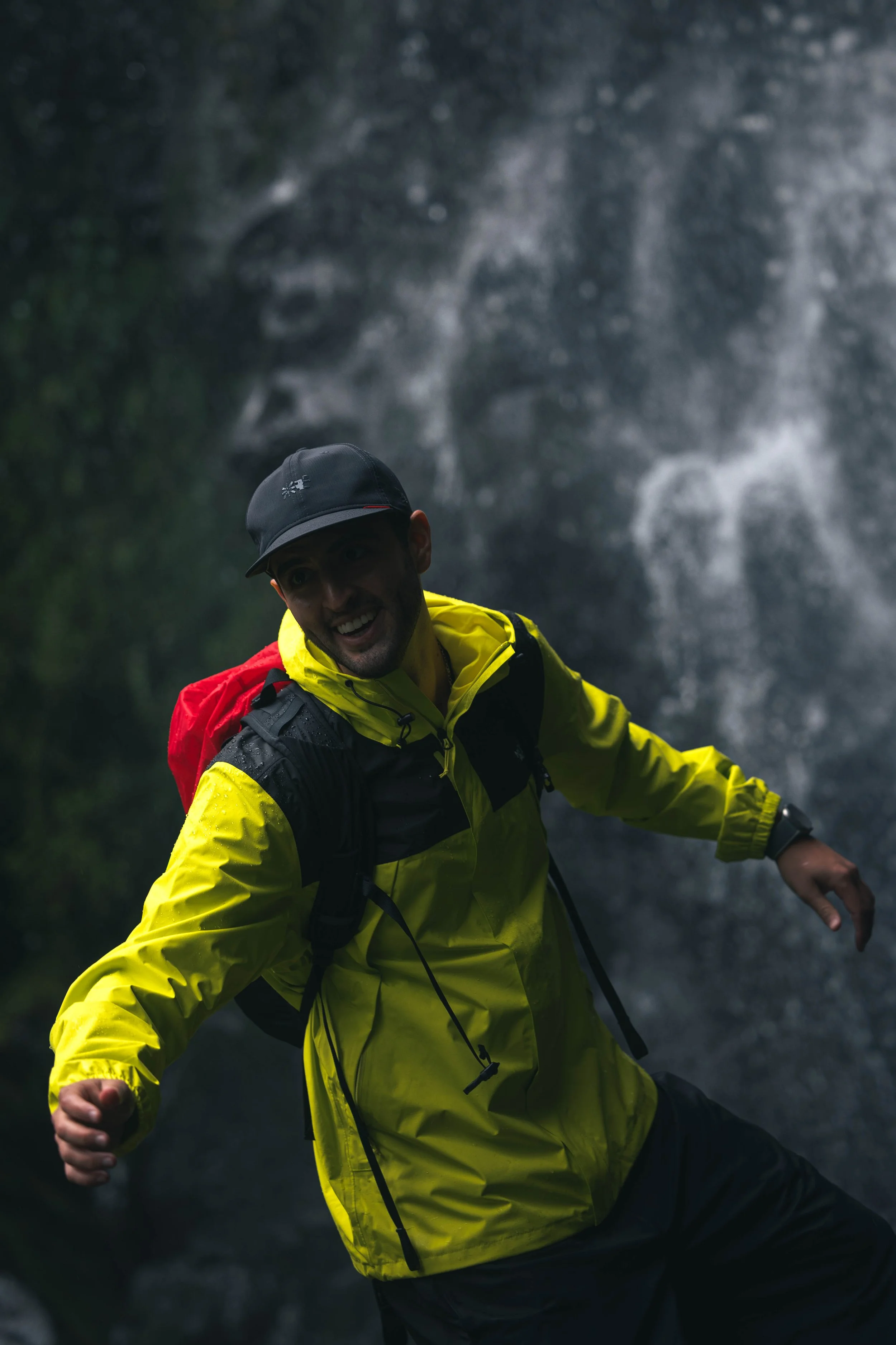 A man wearing a bright yellow rain jacket and a black hat, smiling and enjoying himself outdoors near a waterfall.