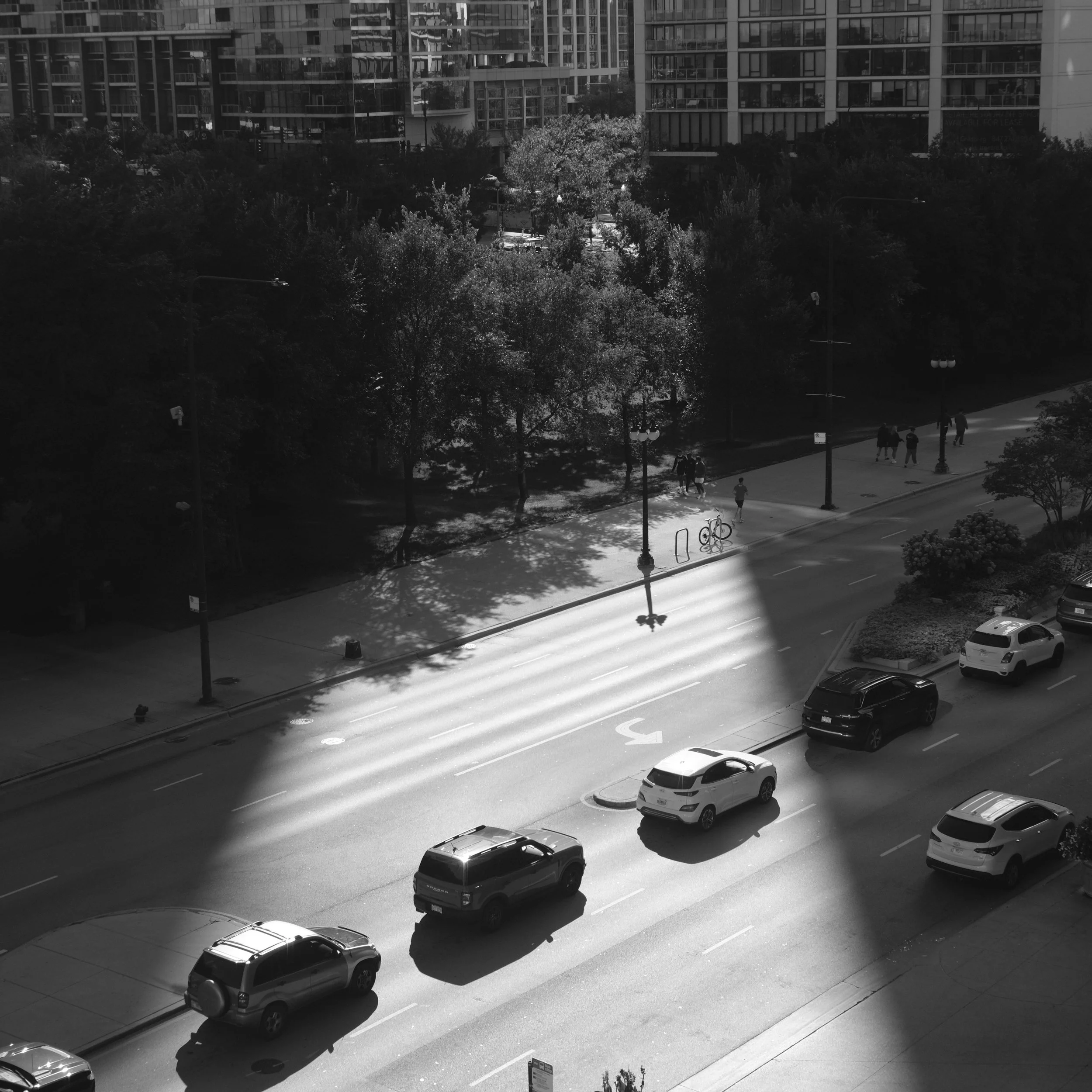 City street with cars, a bicycle, pedestrians on a sidewalk, and trees in a park area, captured in black and white with sunlight casting shadows.