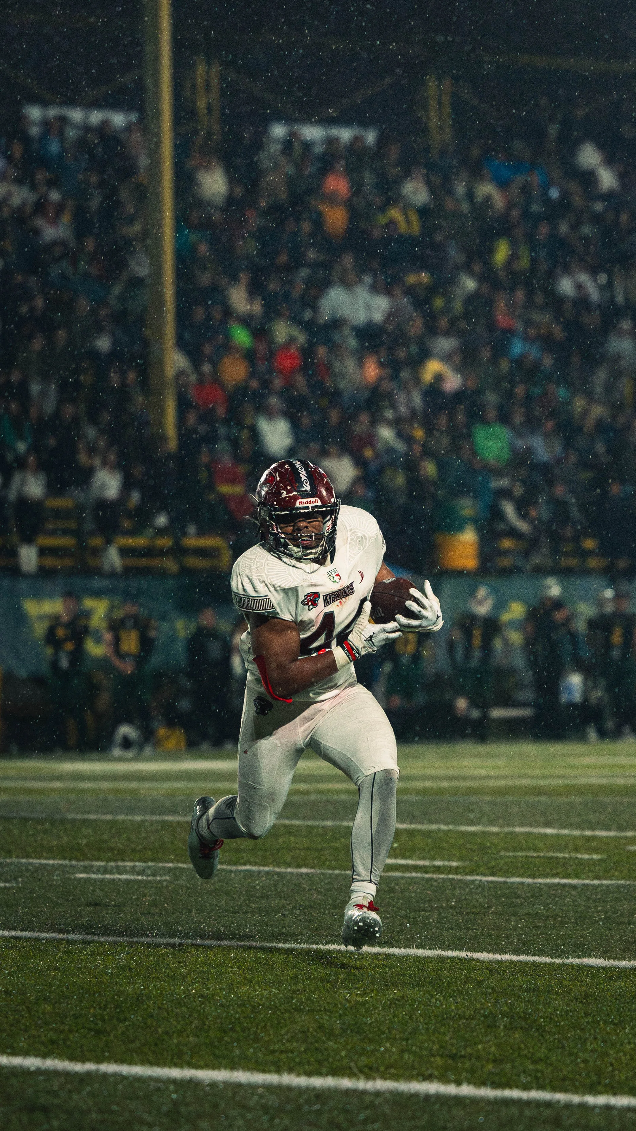 A football player in white uniform running with the ball on a wet field during a game, with a crowd in the background.