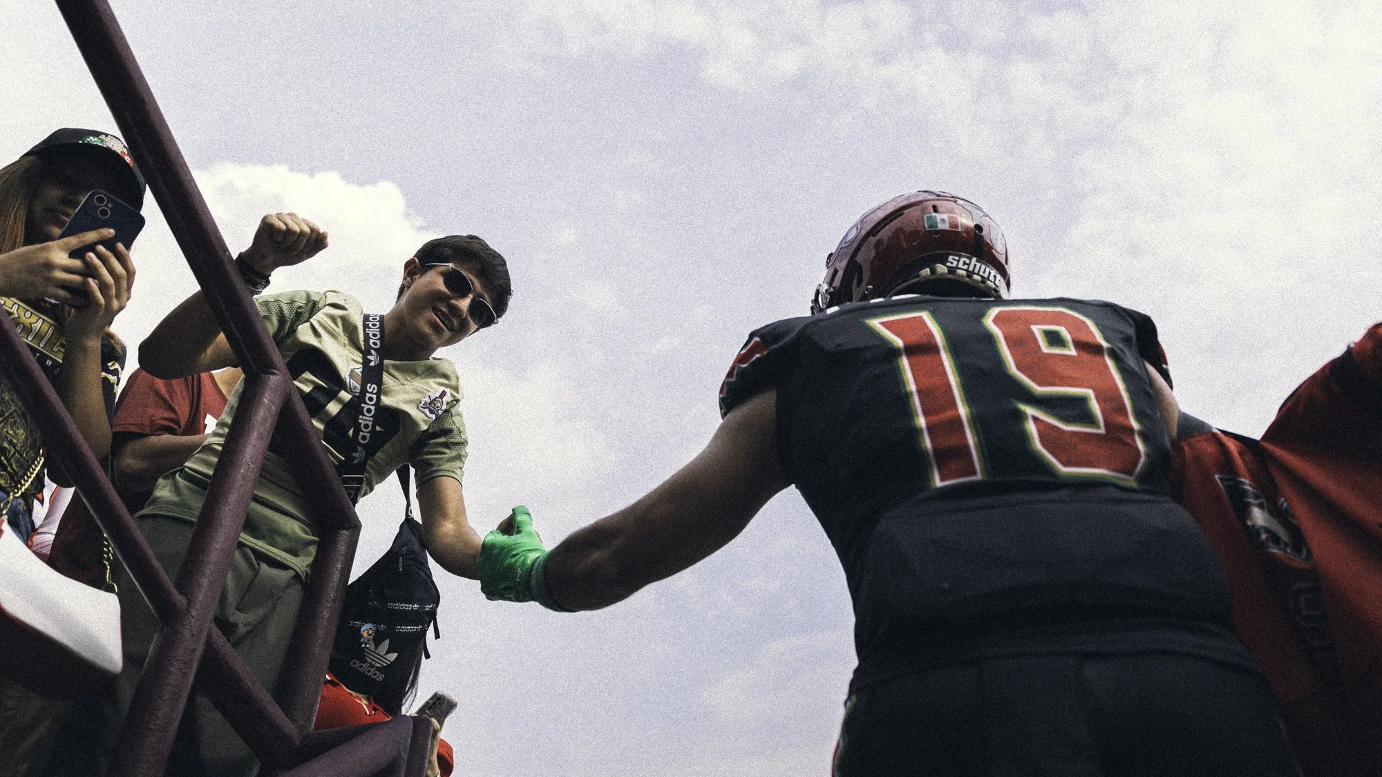 A football player in black and red uniform numbered 19 greeting fans at the sideline, with fans reaching out and giving a fist bump, some taking photos, against a cloudy sky.