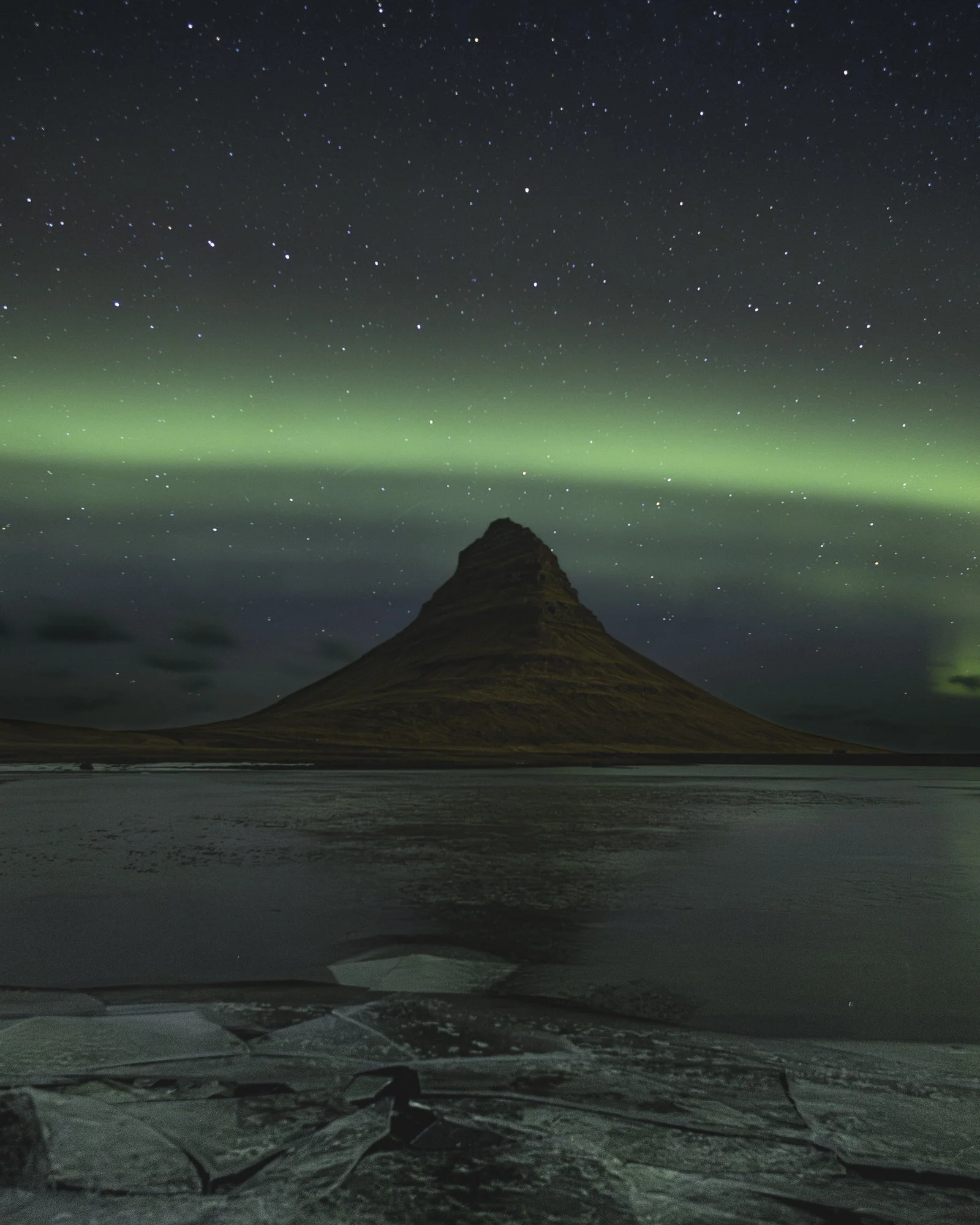 Nighttime view of Kirkjufell mountain in Iceland under the Northern Lights, with stars in the sky and partially frozen water in the foreground.