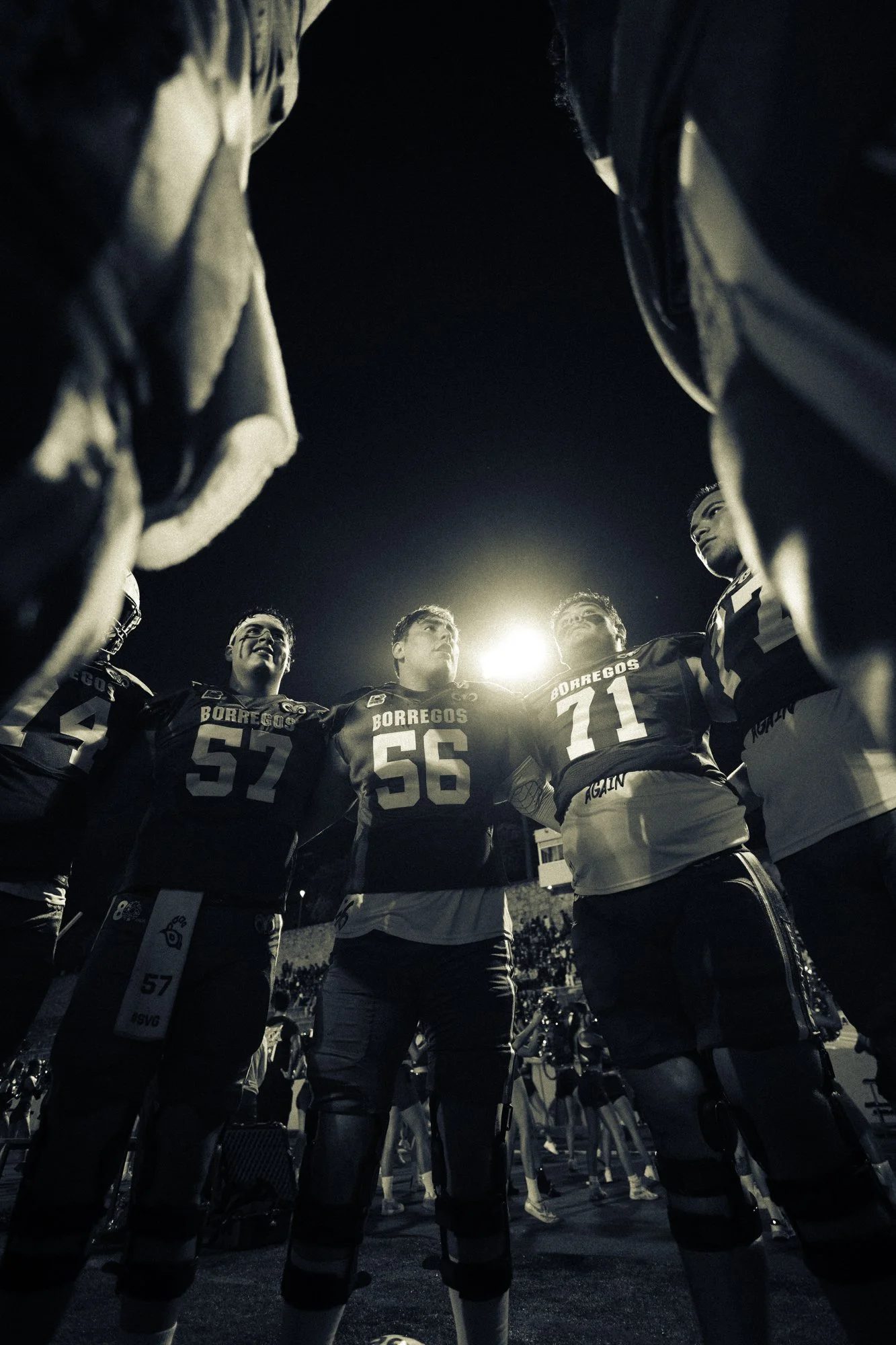 A group of football players in uniform standing in a huddle during a game at night, viewed from below, with bright stadium lights overhead.