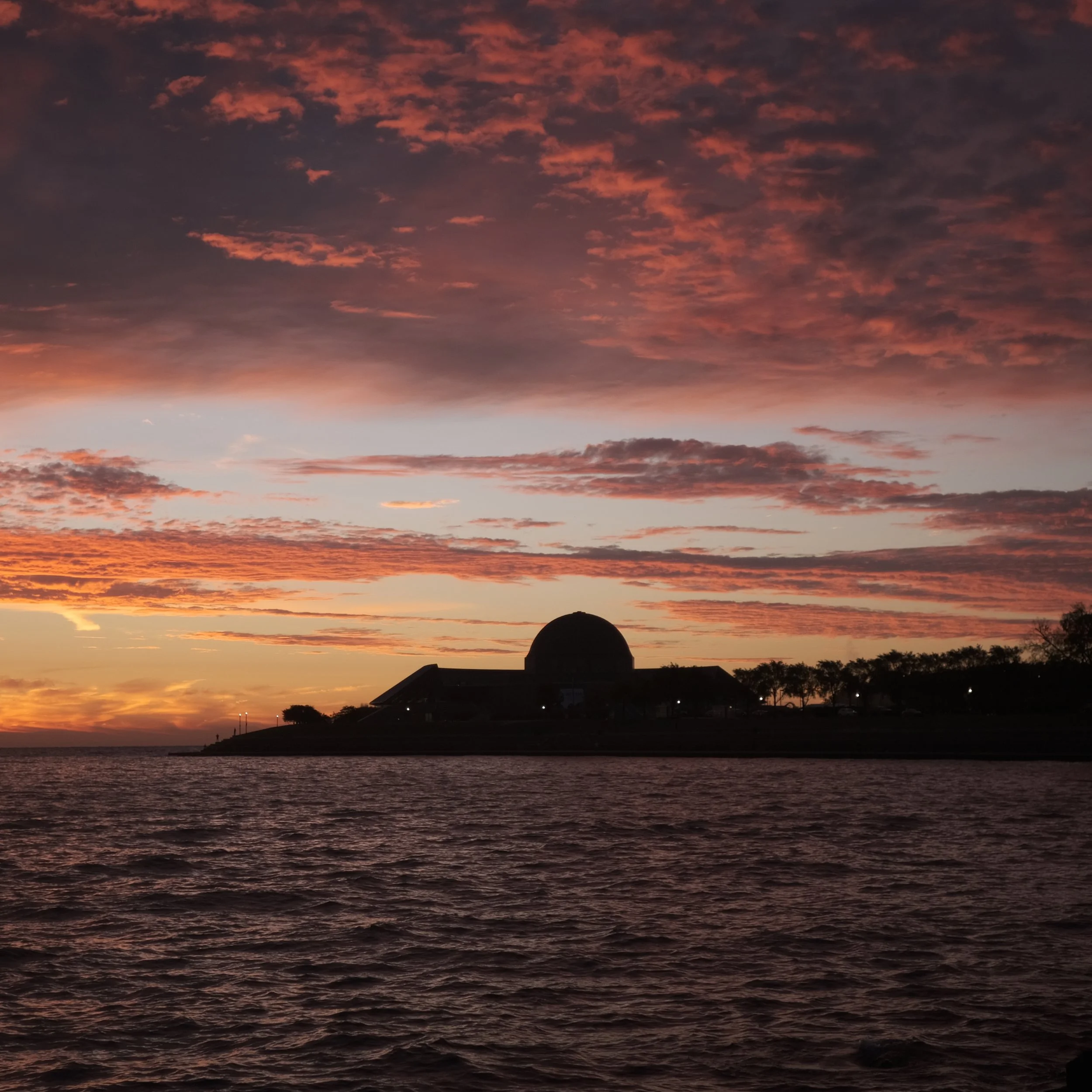 Sunset over a body of water with a silhouetted observatory and trees, colorful sky with orange, pink, and purple clouds.