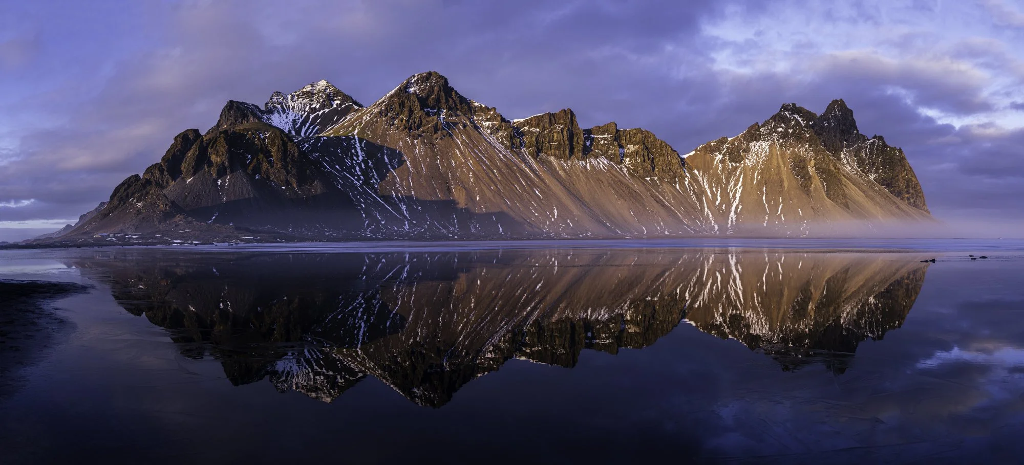Mountain range with snow patches reflected in calm water, under a cloudy sky during sunset.