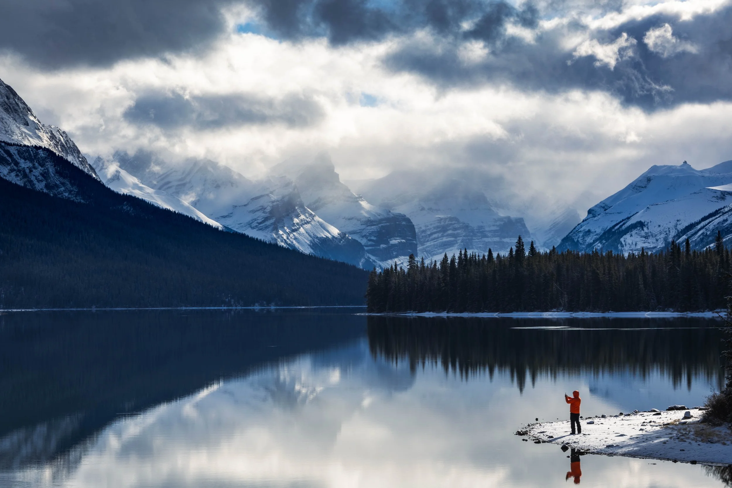 A person in an orange jacket standing on snowy shore taking a photo at a calm mountain lake with snow-covered peaks and pine trees reflected in the water, under cloudy sky.