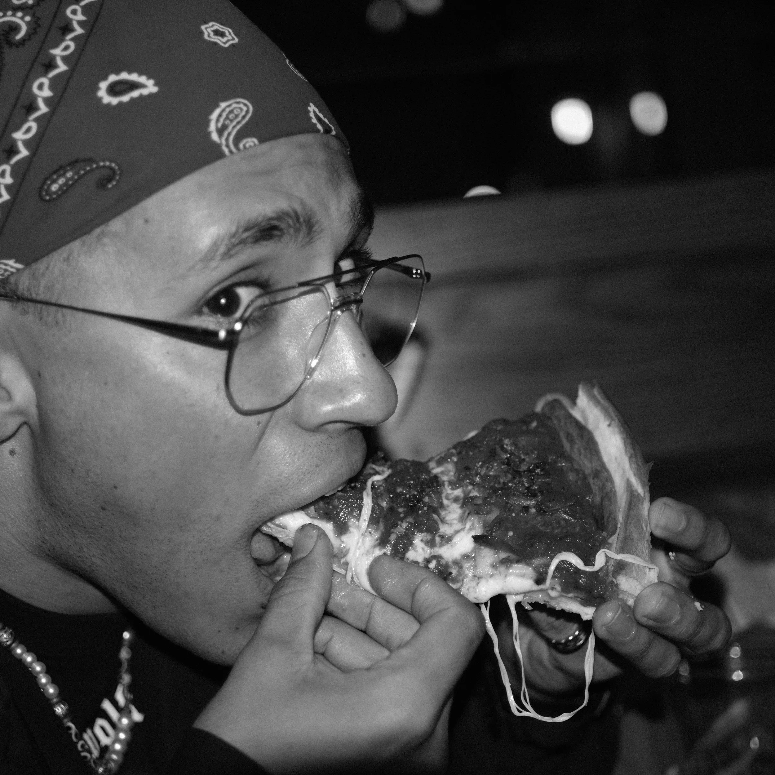 A man wearing glasses and a bandana is eating a slice of pizza.