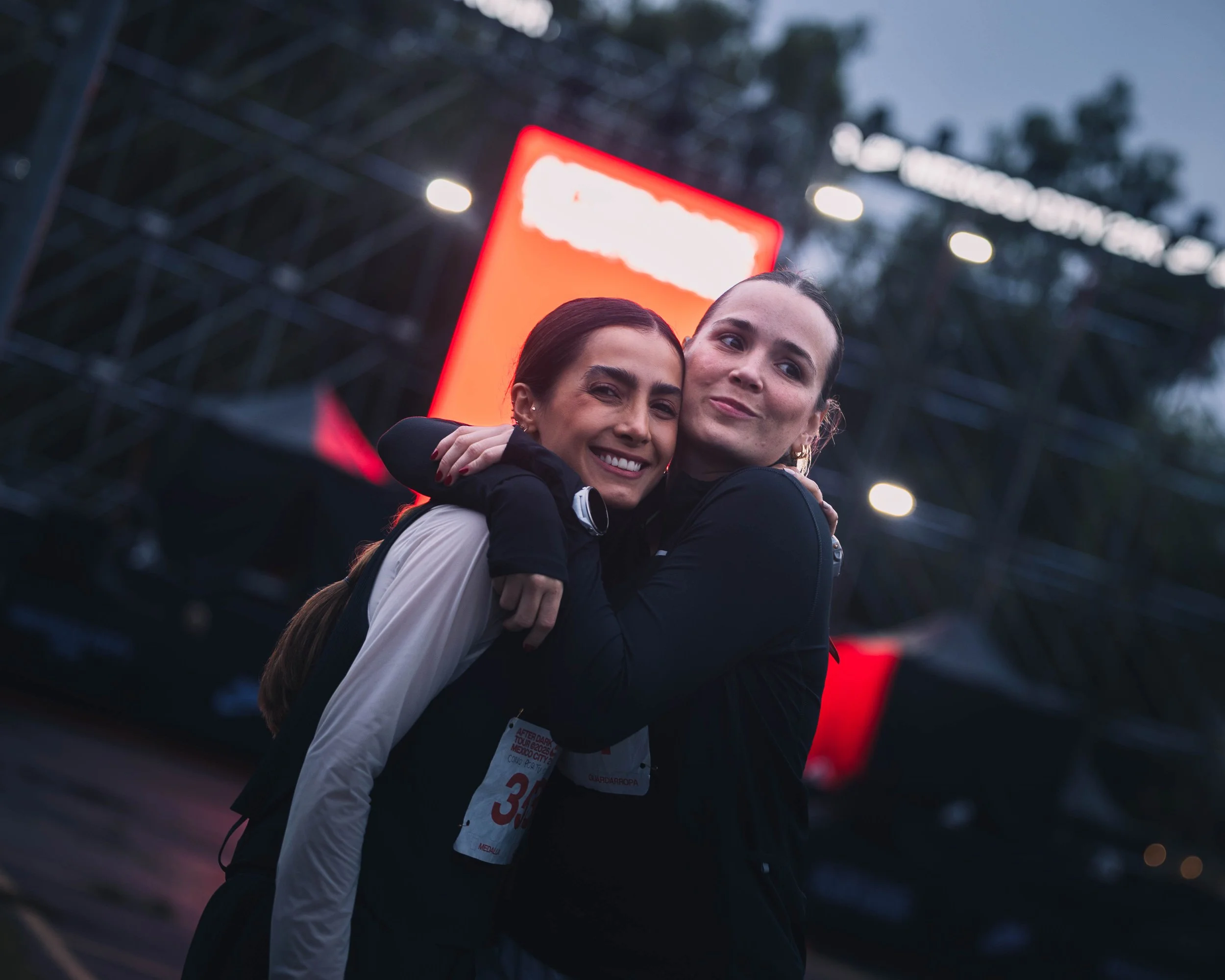 Two women hugging and smiling at an outdoor event during dusk, with a stage and a bright red screen in the background.
