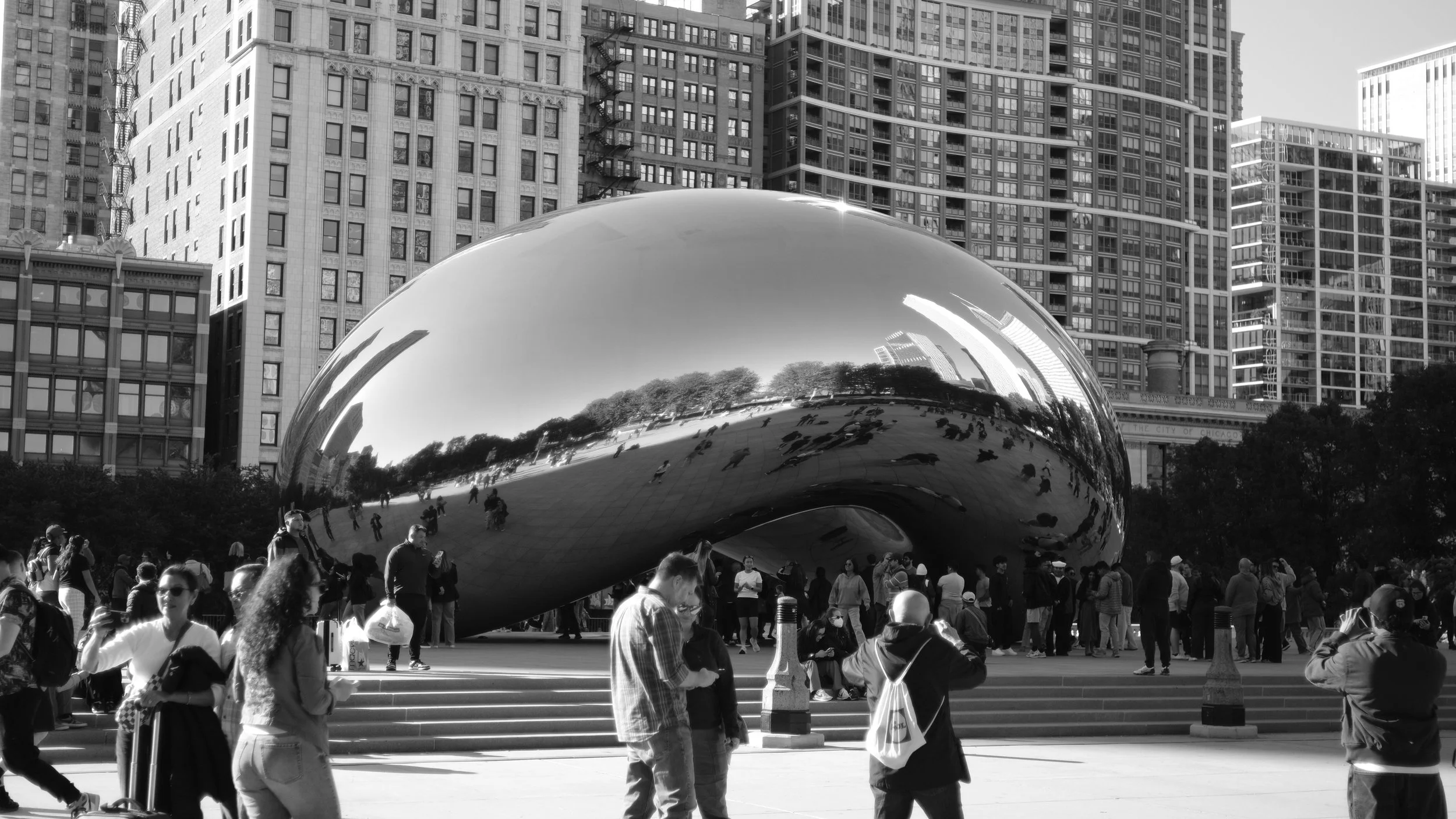 The Cloud Gate sculpture in Millennium Park, Chicago, with people gathered around, surrounded by tall city buildings, in black and white.