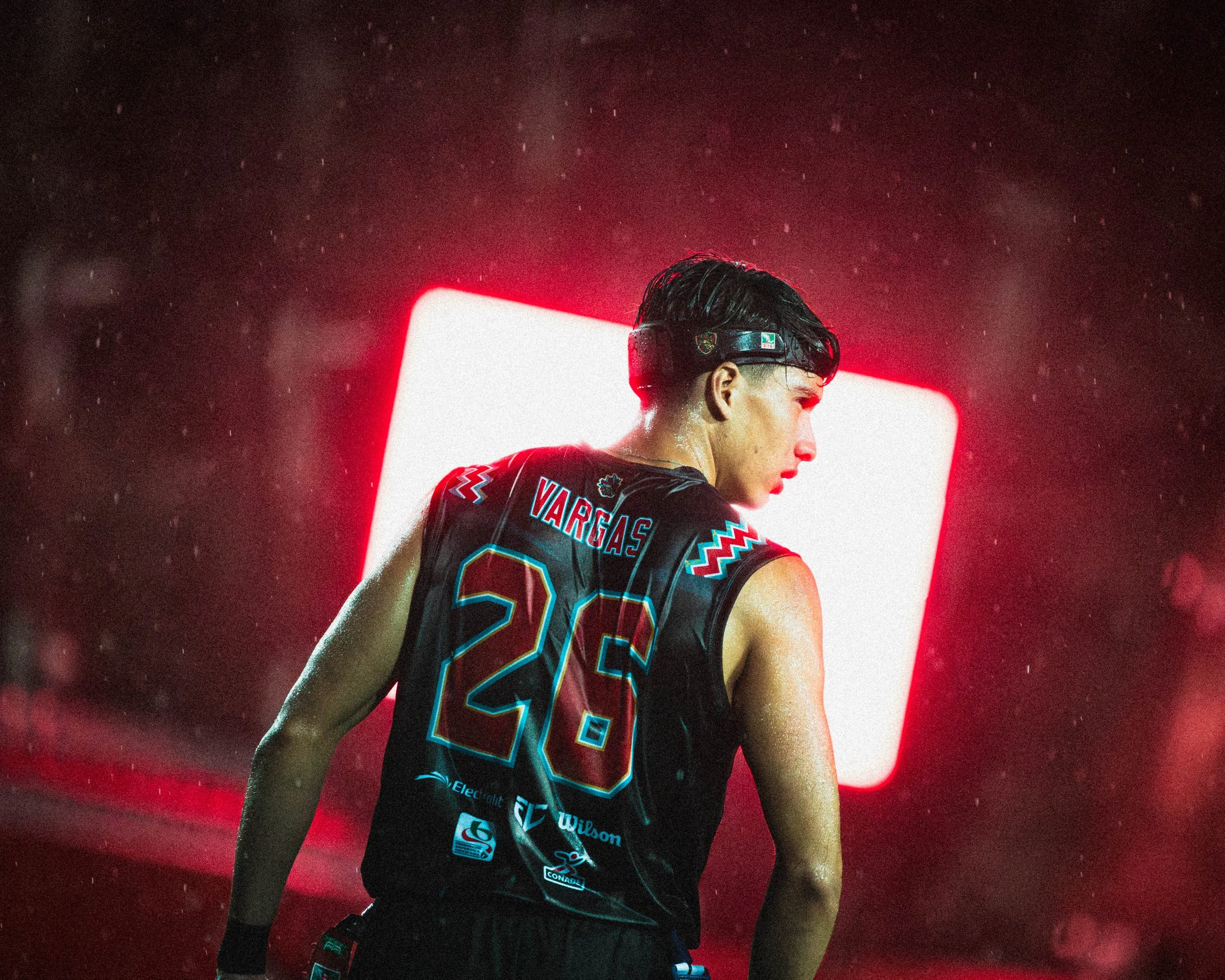 A male athlete wearing a basketball uniform with the number 25 and the name Vargas on the back, standing in a rain-soaked area with a bright white and red light behind him.