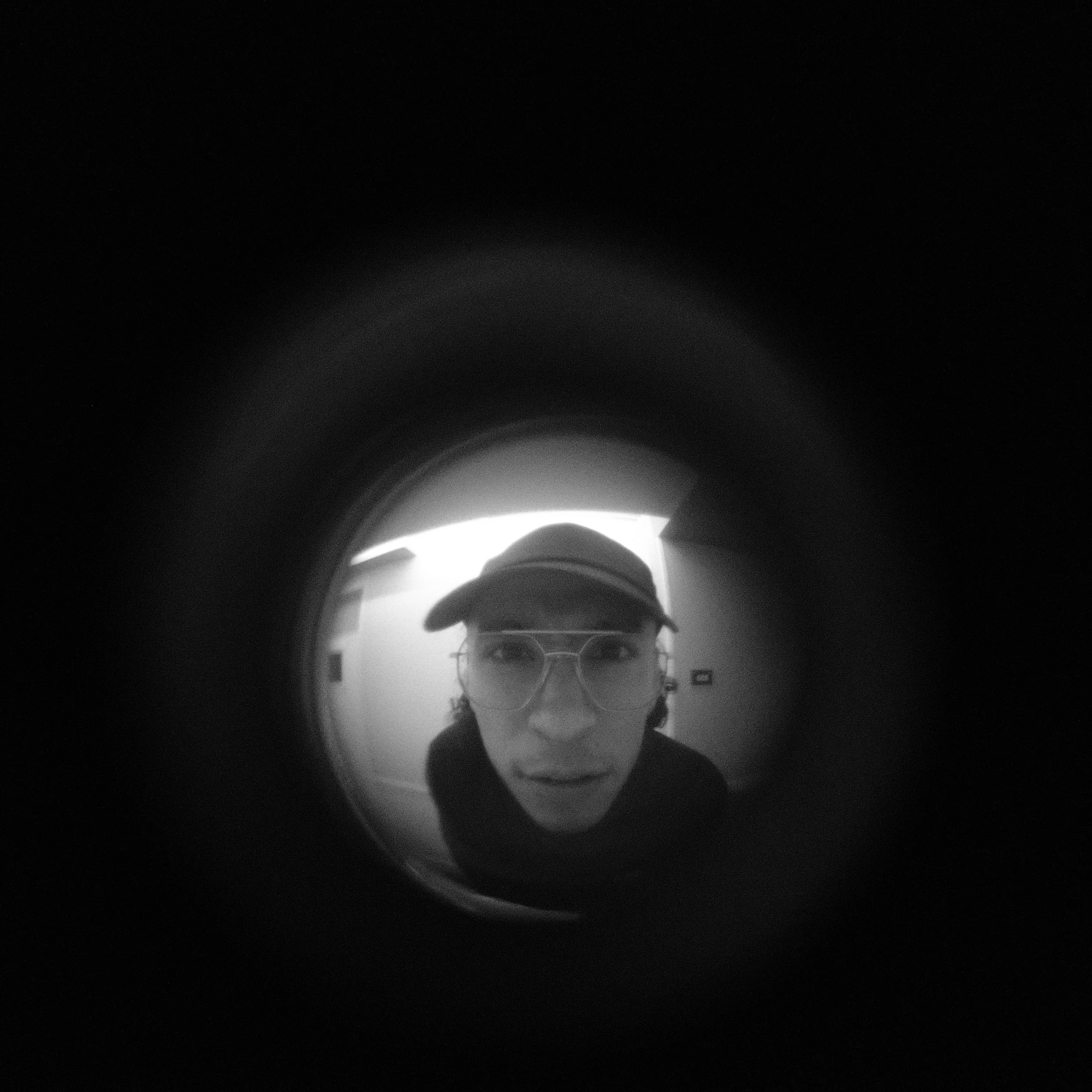 Close-up of a person wearing glasses and a cap, seen through a circular fisheye lens, with a dark surrounding.
