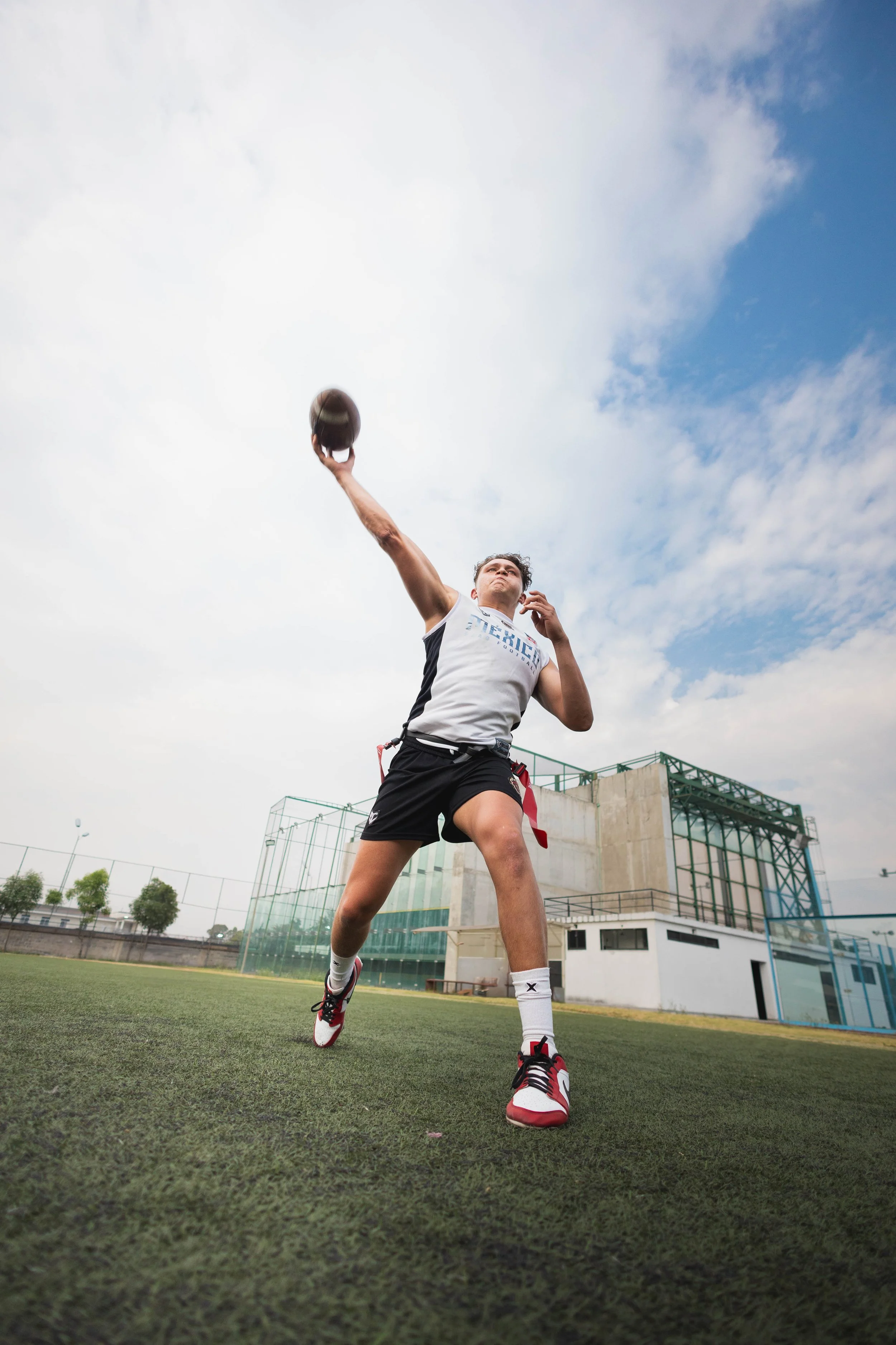 Athlete in sportswear throwing a football on a sports field with a modern building and cloudy sky in the background.