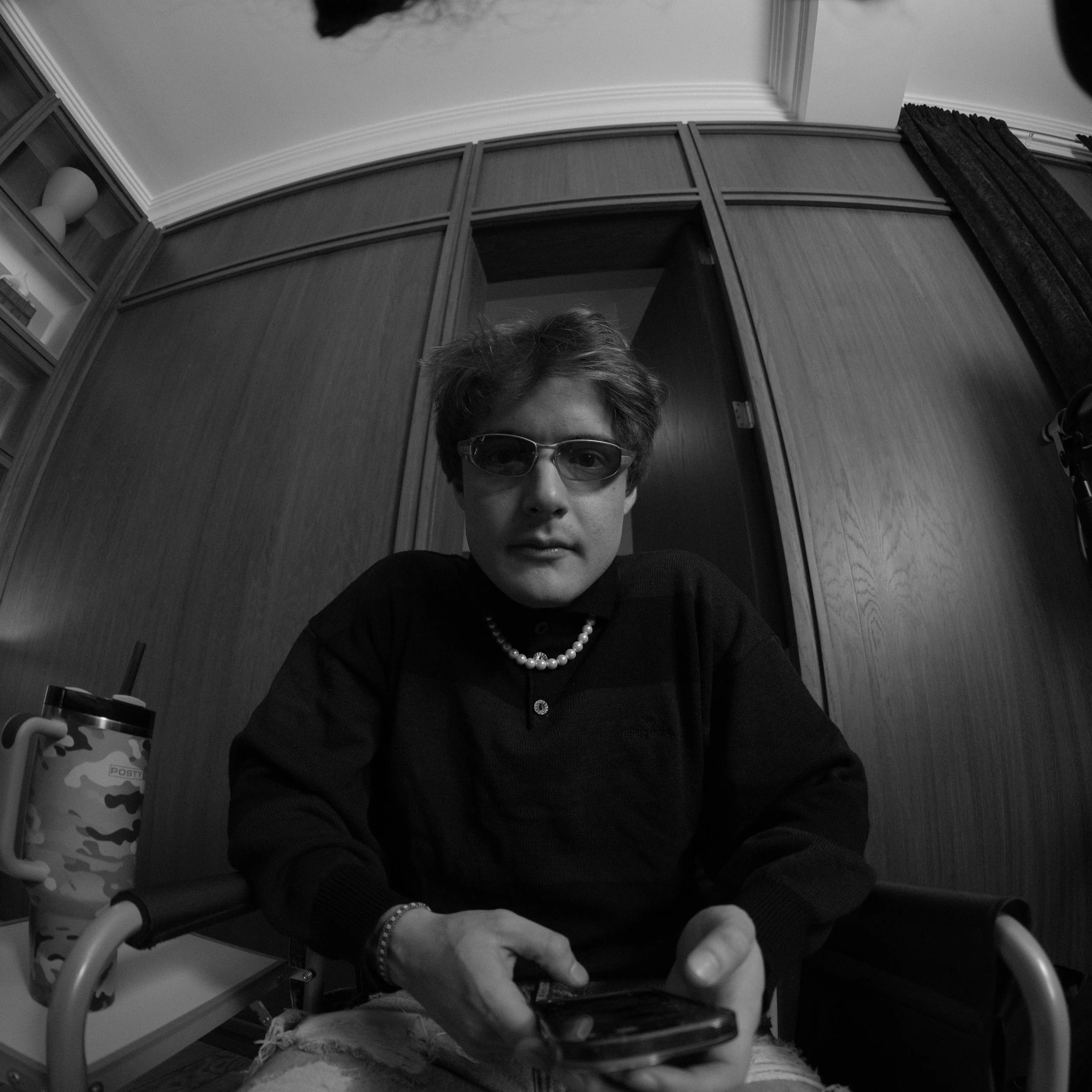 A young person with wavy hair, wearing sunglasses, a pearl necklace, a black sweater, and ripped jeans, sitting indoors and holding a smartphone.