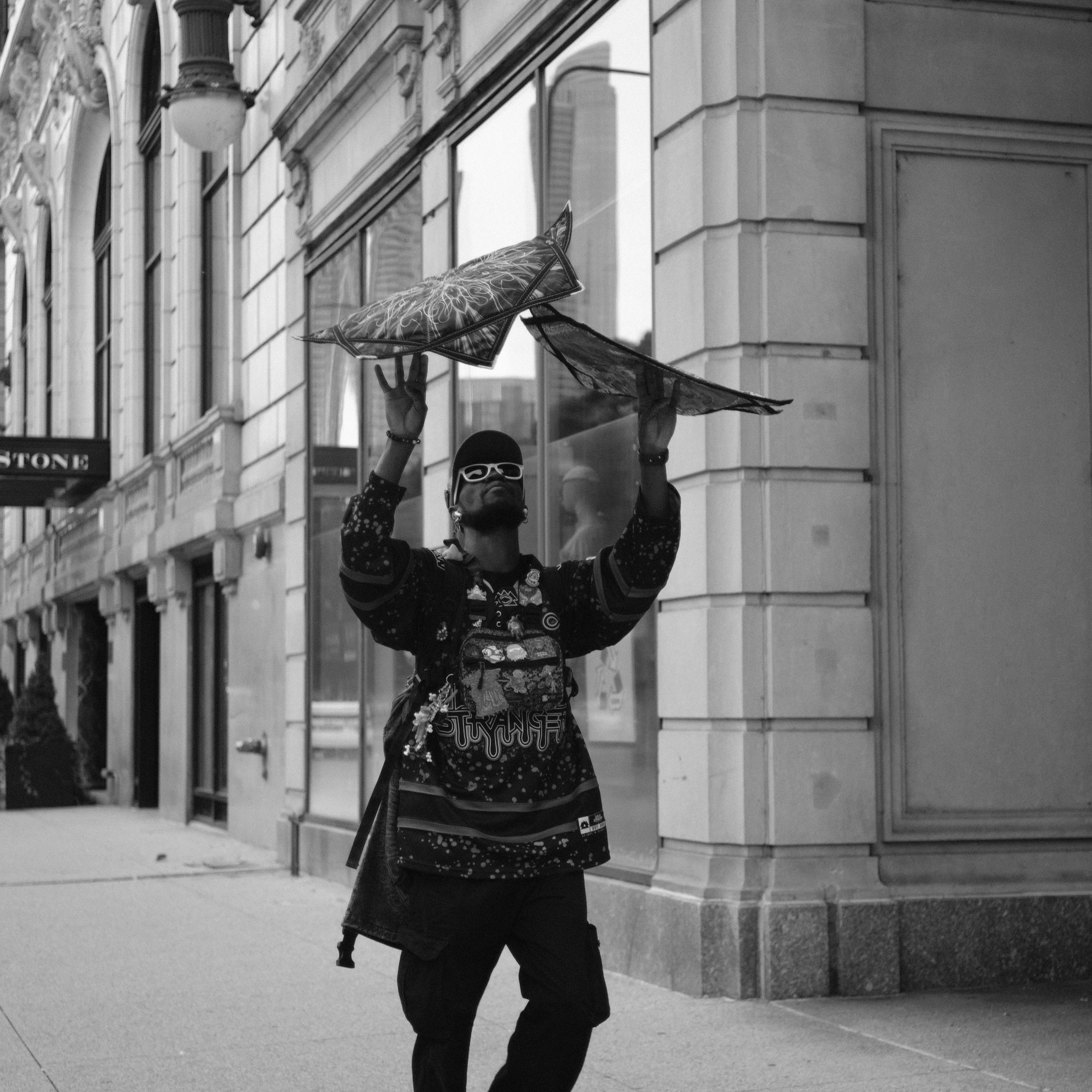 A man wearing sunglasses and a cap stands on a city sidewalk, holding a large umbrella above his head with both hands. The man has a backpack and is dressed in a dark, patterned shirt. The scene is captured in black and white, with tall buildings and
